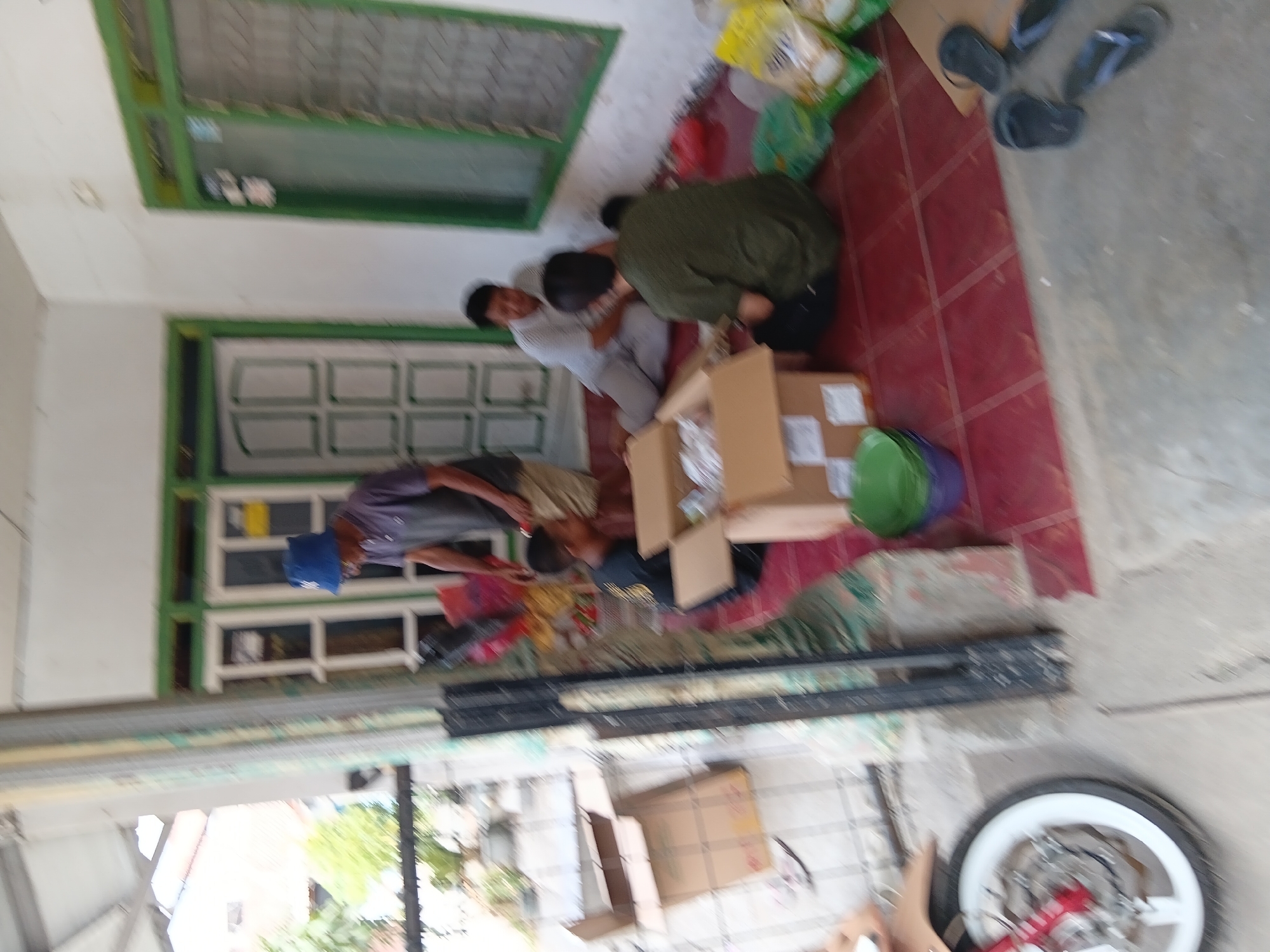 The image depicts an outdoor scene at a building in Tanggul Wetan, Indonesia. Four individuals are present, appearing to be involved in sorting or unpacking items from cardboard boxes. One man, wearing a blue hat and a dark shirt, is standing and appears to be directing or assisting. A younger person in a black shirt is seated and looking down at an open box. Another man, in a light-colored shirt, is seated and leaning towards the box. A fourth individual, wearing a dark green shirt, is also seated and facing away from the camera. Several cardboard boxes, some open and others closed, are scattered around the individuals. Bags of what appear to be rice are visible in the background, along with a motorcycle wheel and a green bucket. The setting suggests a residential or commercial area, with buildings and some greenery visible in the distance. The ground is covered with red tiles.