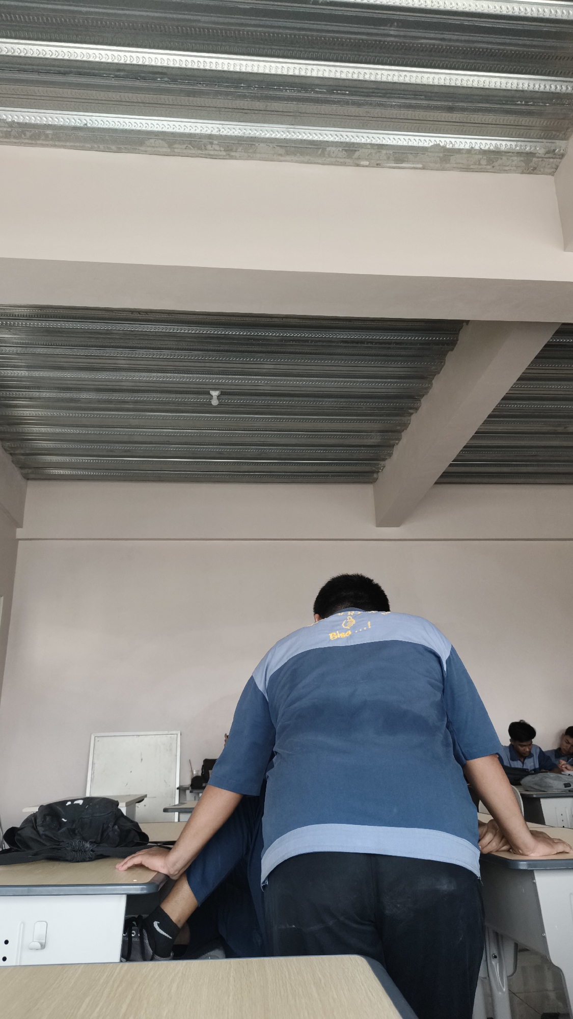 In Katrungan, Indonesia, a classroom is depicted with a focus on the back of a student wearing a blue and light blue t-shirt and black pants. The student is standing, leaning forward with their hands on a desk. Other students are visible in the background, seated at desks. The room features a suspended metal ceiling with visible conduits and a white projector screen on a desk. The overall environment suggests an educational setting.