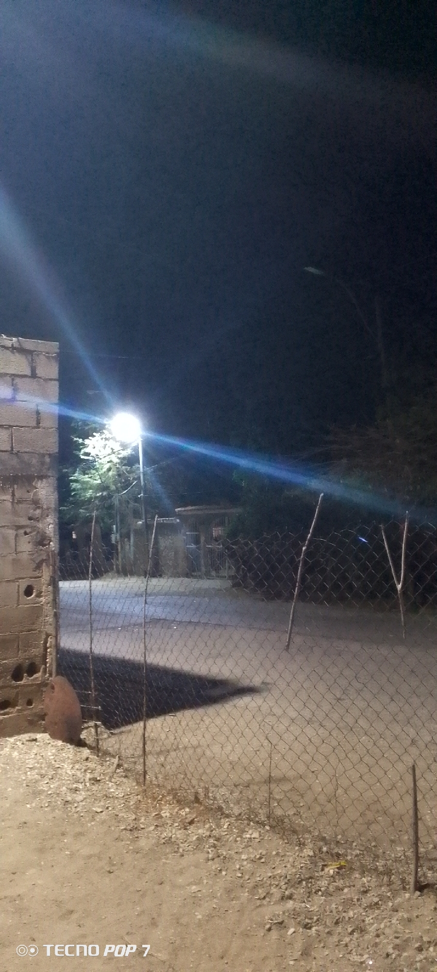 The image, apparently taken at night in Altagracia, Venezuela, depicts a street scene illuminated by a bright street lamp. A cinderblock wall is visible on the left side of the frame. A chain-link fence stretches across the foreground and midground, casting a shadow on the ground. Beyond the fence, the street extends into the distance, with a small structure visible on the right. The sky is dark. The ground is dirt or gravel. A "Tecno Pop 7" watermark is present.