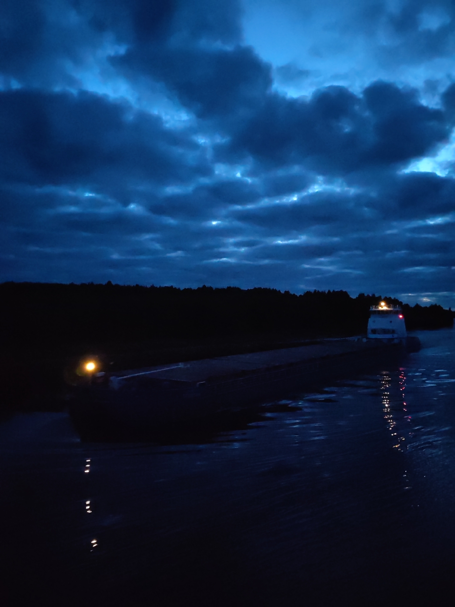 A nighttime scene with a large, presumably nautical, vessel or ferry traveling on water. The vessel is illuminated with lights, including a prominent light on its upper structure. A shoreline or landmass with trees is visible in the middle ground. The sky is filled with dark, ominous clouds. Reflections of the vessel's lights are visible in the water. There is no indication of the location being Uzhla, Russia, or any human presence. The scene depicts the movement of the vessel in an environment dominated by water and a cloudy night sky.