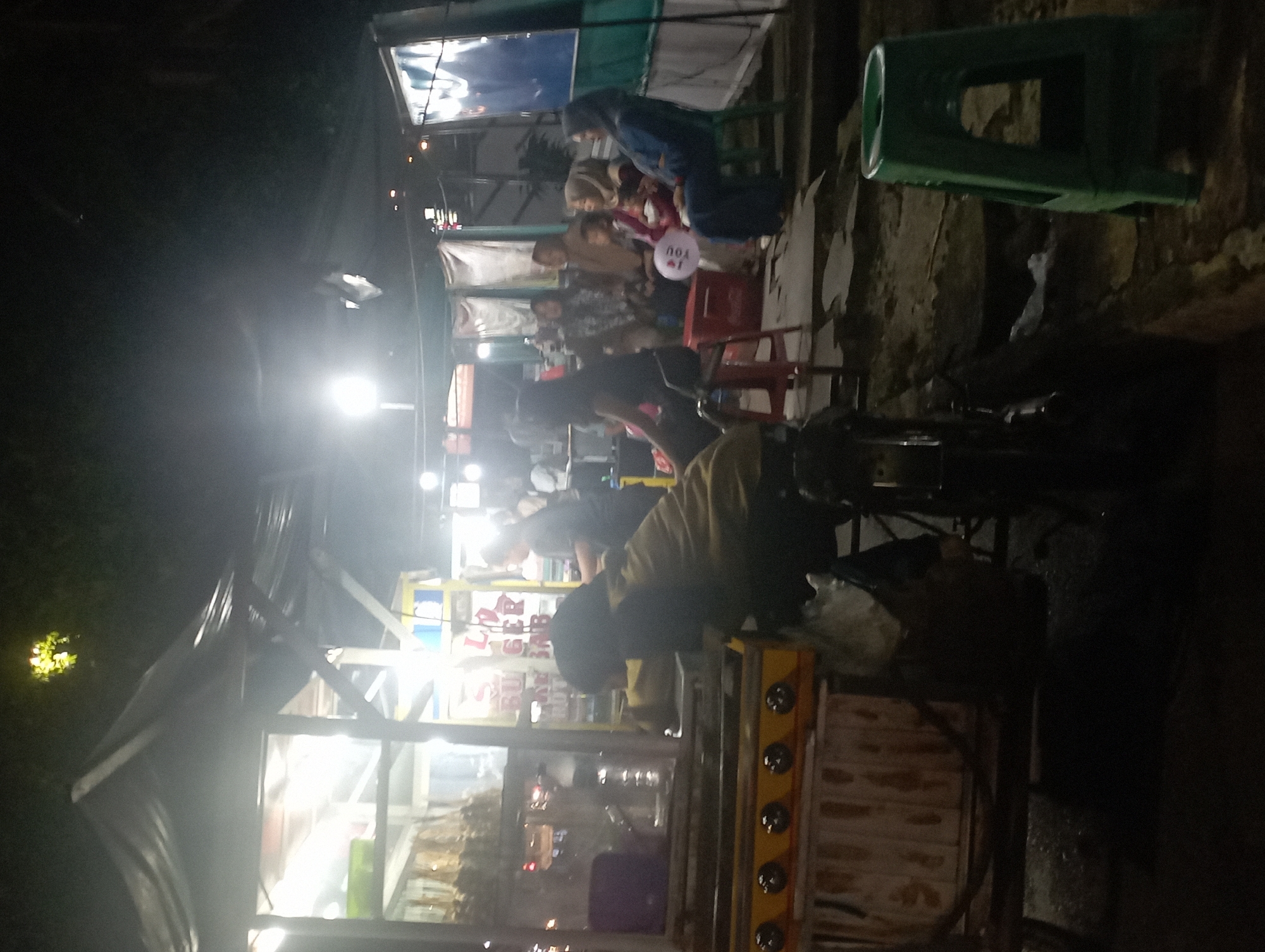 At night, an outdoor food stall operates in an urban setting. The stall features a metal and wood cart with a yellow front panel, equipped with burners. Inside the stall, illuminated by bright lights, various food items are displayed, including what appears to be fried snacks. A person in a tan jacket is bent over the cart, seemingly preparing food. Behind the stall, a group of people are seated, some of them wearing headscarves, suggesting a casual gathering. A child is present, and a white balloon with text is visible. The ground is paved with concrete and dirt. The scene is dimly lit, with artificial lighting from the stall and overhead lamps casting shadows.