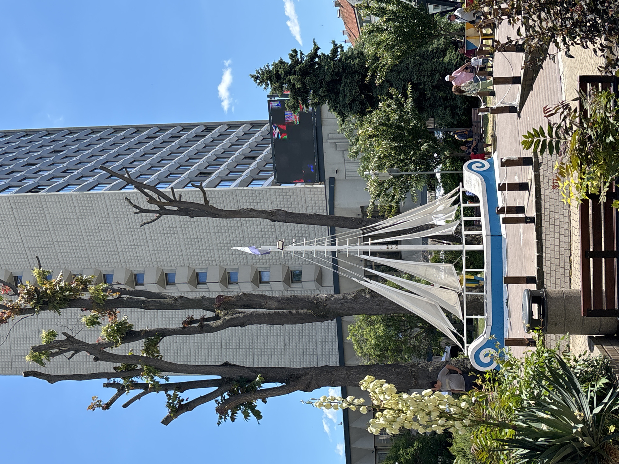 This image depicts a sunny day in Burgas, Bulgaria, likely in a public square or park. In the foreground, a decorative model of a sailing boat, painted blue and white, is prominent. The boat sits on a wooden deck surrounded by low wooden posts and rope railings. Lush green plants, including a tall plant with white flower clusters, are to the left of the boat, and other foliage lines the foreground.
Behind the boat and greenery, several tall, bare trees stand. In the background, a large, modern, light-colored building with many windows dominates the skyline. To the right of this building, a large, dark digital billboard displays colorful abstract graphics. More trees, including a dark green evergreen, are visible between the building and the billboard.
Several people are scattered throughout the scene, appearing to be enjoying the outdoor space. A man in a grey t-shirt stands near the boat, leaning against one of the trees. Further back, near some benches, several individuals are seen sitting and standing, interacting in small groups. The overall atmosphere suggests a pleasant, public gathering place.