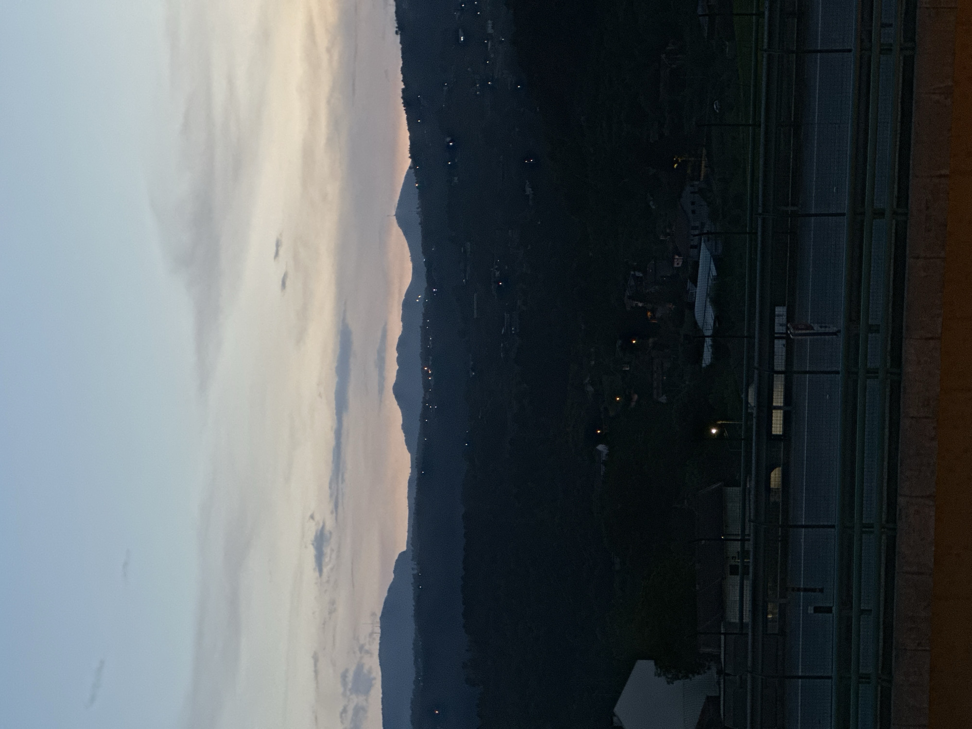 The image presents a view from an elevated position, likely a building in Vila Real, Portugal, facing a mountainous landscape at dusk. The sky is a mix of light blues and soft grays with scattered clouds. Silhouetted mountains with sparse light points suggesting dwellings or streetlights are visible in the middle ground. The foreground features a section of a building, including a window and partial façade, overlooking a road with some fencing. There are a few scattered lights from buildings or vehicles. The overall scene captures a transition from day to night in a semi-urban setting.