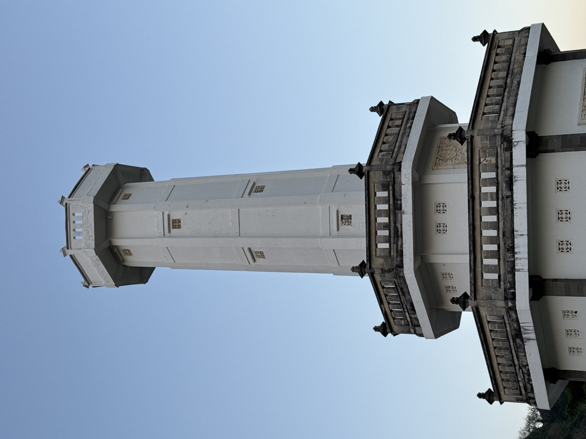 A white octagonal tower stands vertically against a clear, light blue sky. The tower features multiple tiers, beginning with a broader base that gradually tapers upwards. The lower section includes at least two visible octagonal balconies, each with dark grey or brown stone balustrades featuring both vertical slats and solid rectangular panels, topped by decorative finials at the corners. The walls of the tower are primarily white, displaying intricate square cutout patterns, reminiscent of jali work, in a grid arrangement on the lower levels. One of the lower balcony levels also shows a carved relief panel. The main shaft of the tower is an octagonal column, also white, with subtle horizontal and vertical linear details. Further up, it features more of the square decorative cutouts, appearing in a lighter, golden hue within recessed panels. The uppermost section of the tower is narrower, with an octagonal platform topped by a crenellated-like parapet with vertical openings. The overall construction appears to be concrete or stucco, with darker stone elements. There is minor discoloration and weathering visible on the white surfaces, particularly under the darker stone sections. A small portion of green foliage and a dark hillside are visible on the far left edge of the image, at the base of the structure. No individuals or active elements are present in the scene. This structure is located in Kuta Selatan, Indonesia.