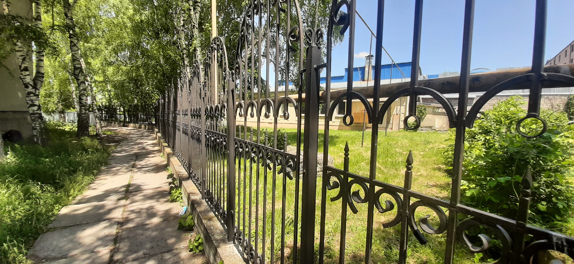 A metal fence with ornate scrollwork and pointed finials borders a paved walkway lined with birch trees on the left. Beyond the fence, a grassy area is visible, with a light-colored building featuring arched windows and a blue-roofed structure with a tall chimney in the background. The scene appears to be a section of the grounds of a sanatorium or recreational facility in Yessentukskaya, Russia, under a clear blue sky.