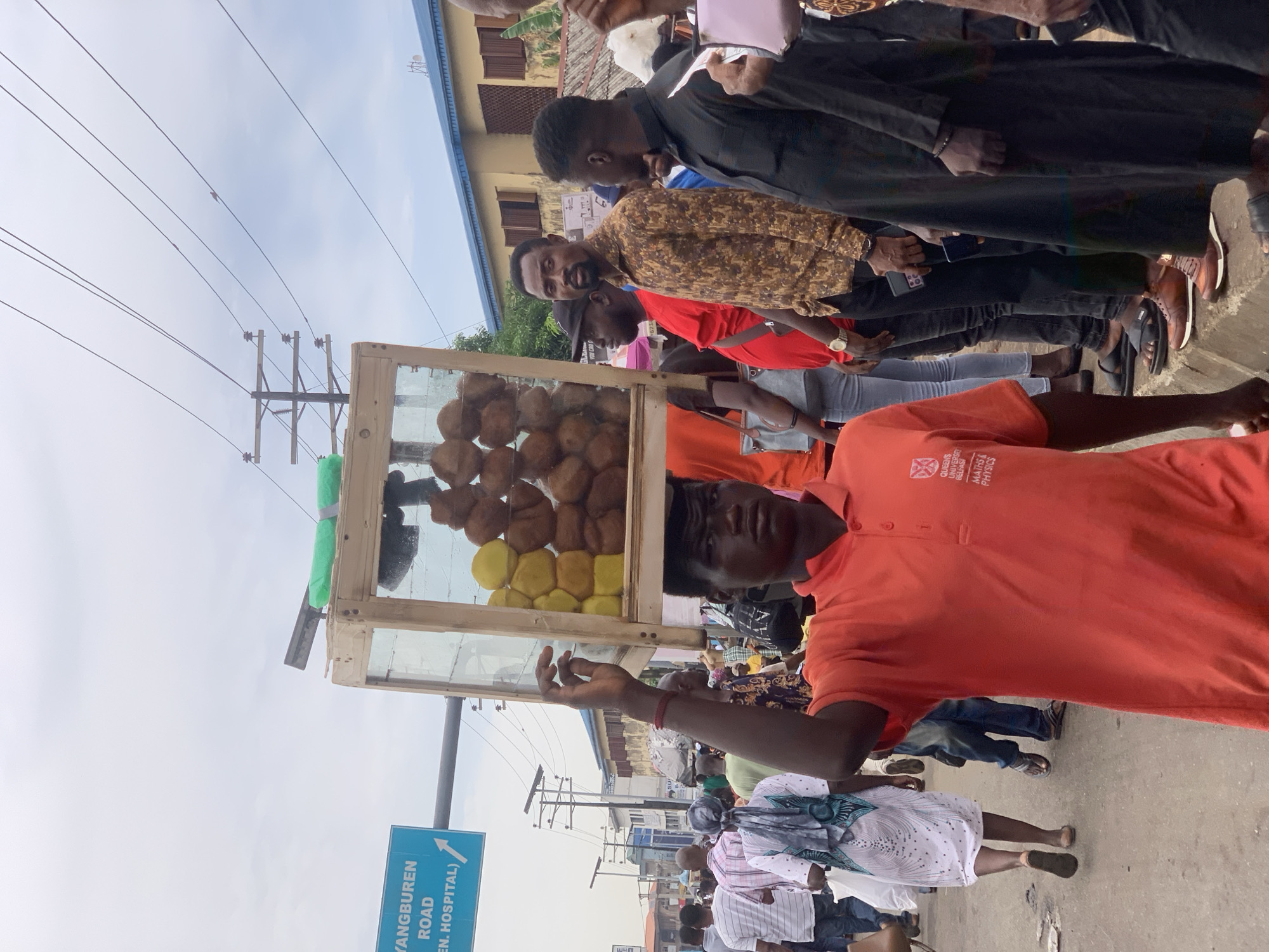 A street vendor in Ikorodu, Nigeria, stands in a crowded area, selling what appears to be fried dough. He is wearing an orange shirt bearing the logo of Queen's University Belfast, specifically the department of Maths & Physics. The vendor's goods are displayed in a wooden and glass case balanced on his head. He is surrounded by people, some of whom seem to be customers. A street sign visible in the background reads "Yangburen Road." Overhead power lines are visible, and the sky is overcast.