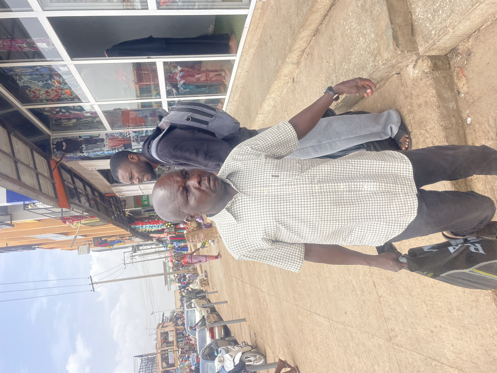 The image shows two men standing on a sidewalk in Jos, Nigeria. The man closest to the camera is older, with a bald head, wearing a checkered short-sleeved shirt, dark pants, and sandals. He is holding a black bag. Behind him is a younger man with a beard, wearing a dark hoodie and gray sweatpants. He has a backpack. They are standing in front of a building with a glass facade that reflects the street scene. The street is lined with cars and people, suggesting a busy commercial area. Overhead power lines are visible. The scene is captured in daylight.