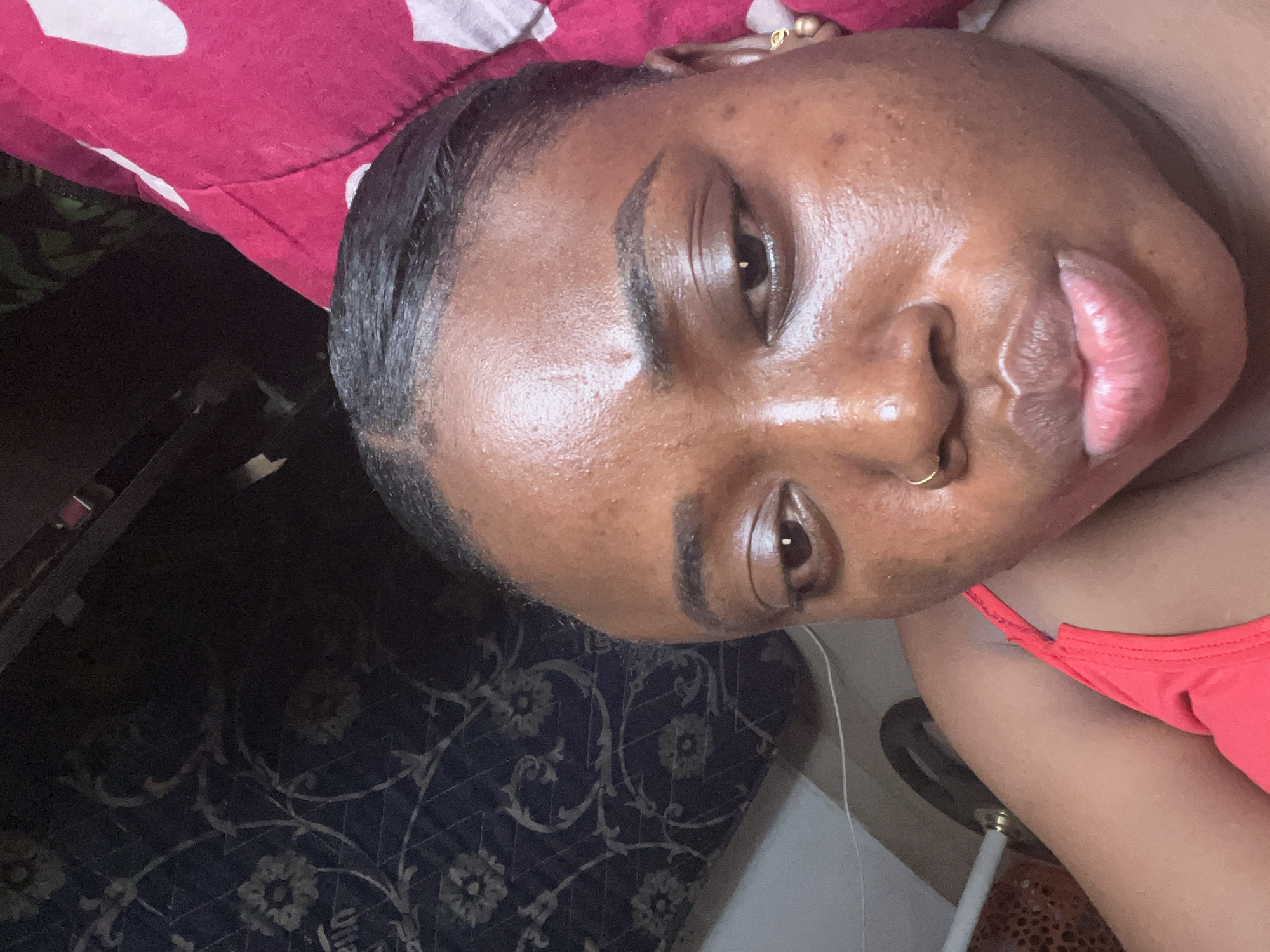 A close-up selfie of a woman with dark skin, brown eyes, and black hair is shown. She has a nose ring and earrings. She is wearing a red top. The background includes a red and white patterned surface and a dark blue surface with a floral pattern. A white cord is visible near her right arm. No evidence or reference points suggest the location is Abuja, Nigeria.