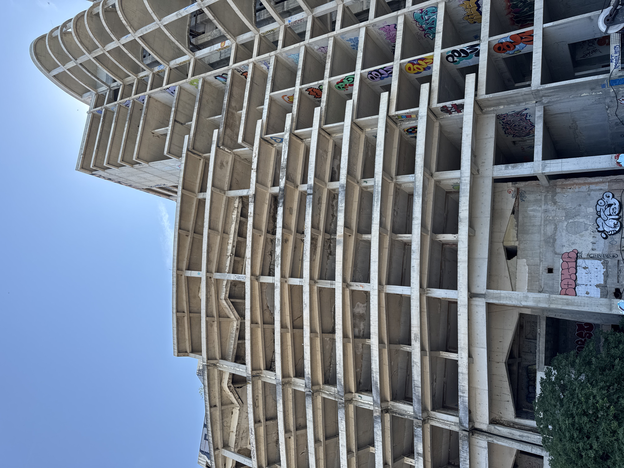 The image shows an unfinished, multi-story concrete structure in Perazića Do, Montenegro. The building's exterior consists of repeating horizontal slabs and vertical supports, creating numerous open floors and balconies. The raw concrete surfaces are heavily covered with various styles of graffiti, including tags, bubble letters, and more intricate designs in a range of colors. The sky is clear and blue. There is a dense green tree on the left side of the frame, partially obscuring the lower levels of the building. The scene suggests abandonment or a prolonged construction halt.