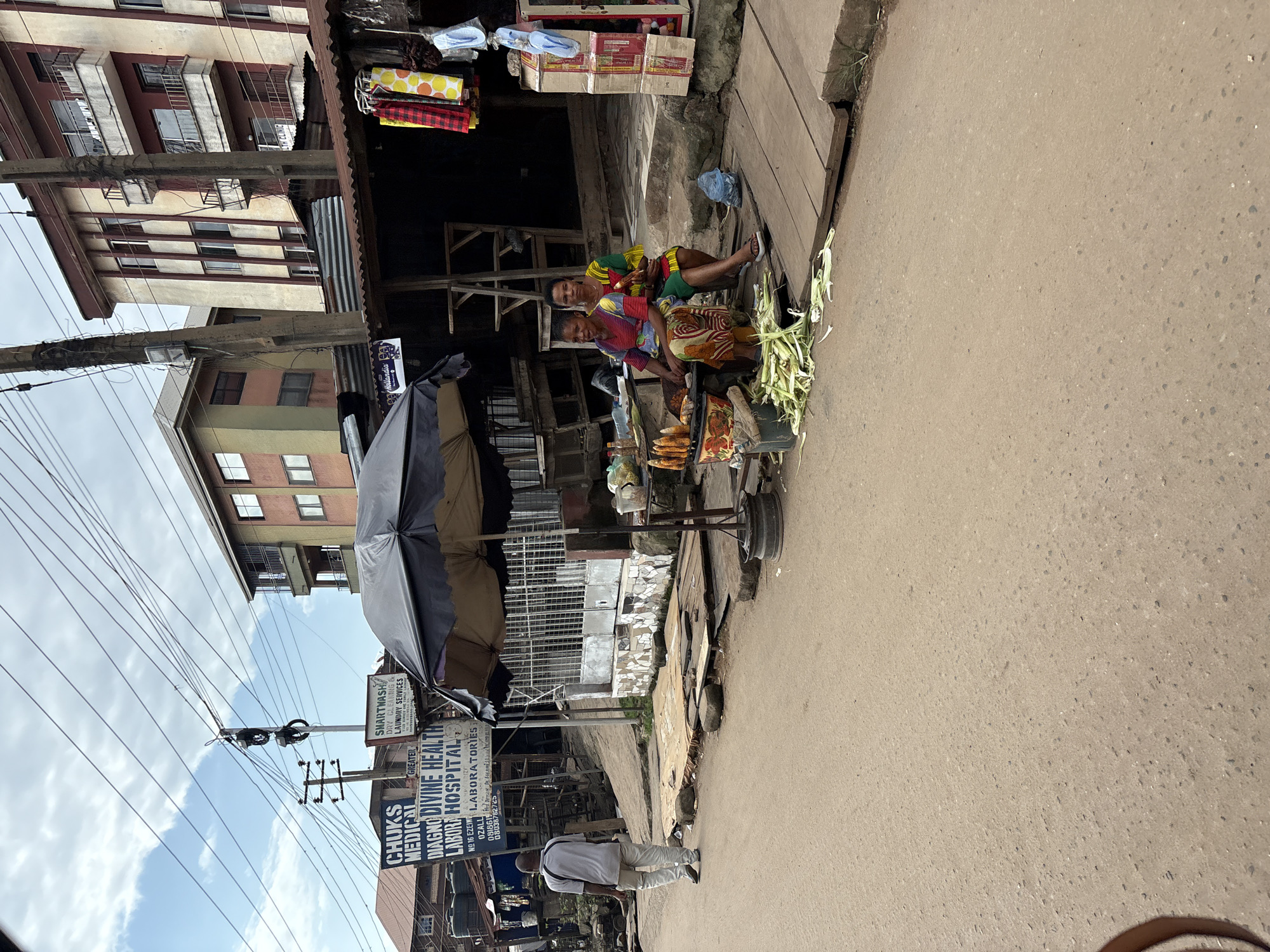 This is a street scene in Onitsha, Nigeria, featuring a roadside market. In the foreground, a man in a grey t-shirt and light-colored trousers walks away from the camera on a paved road. To his left, a collection of signs advertises medical services, including "CHUK'S MEDICAL," "GREATER DIAGNO DIVINE HEALTH LABOR HOSPITAL," and "LABORATORIES." To the right of the road, two women are seated at a small market stall. One woman, wearing a colorful patterned dress, is holding corn on the cob. The other woman, in a green and yellow dress, is seated beside her. In front of them, a pile of corn husks lies on the ground, and several ears of corn are displayed on a grill. Behind the stall, a dark awning covers merchandise, and clothing items are hanging on display. In the background, multi-story buildings with balconies line the street, and electrical wires crisscross overhead. The sky is overcast.