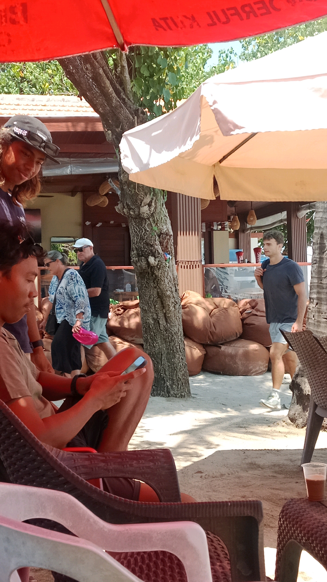 The image depicts an outdoor setting, likely a beach bar or cafe, in Kuta, Indonesia, given the visible text on the red umbrella. In the foreground, a person is seated in a chair, looking at a mobile phone. Their legs are crossed, and they are wearing shorts. To their left, another person is partially visible, wearing a dark blue t-shirt.
Behind the seated individual, a couple walks past. The woman on the left wears a patterned blue and white blouse and dark pants, carrying a pink object in her hand. The man beside her wears a white cap, sunglasses, a dark t-shirt, and light-colored shorts.
Further in the background, a man in a dark t-shirt and light shorts walks away from the camera, carrying a backpack. He appears to be walking towards a building with a counter, possibly a bar or restaurant. The ground is sandy, characteristic of a beach environment. Large brown beanbag chairs are scattered around the area. The scene is shaded by a red umbrella and a cream-colored umbrella. Tropical trees and foliage are visible in the background, suggesting a warm climate.
