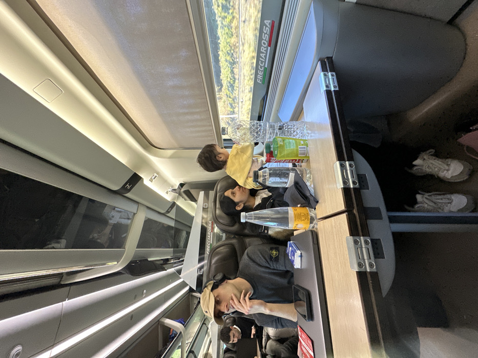 This image captures the interior of a Frecciarossa high-speed train in Italy, traveling through an area near Civita Castellana. The train carriage features modern design with overhead luggage racks, strip lighting, and large windows. Outside, blurred green vegetation suggests motion.

In the foreground, a wooden foldable table is laden with items. On the left side, an open grey laptop, a black smartphone, a blue and white tissue pack, and two red cards are visible. On the right, various beverages are present: a clear plastic bottle with a yellow label, a dark cloth or pouch, a partially obscured clear bottle, a red Coca-Cola can, a green plastic bottle, and a large textured clear plastic water bottle. Beneath the table, two pairs of light-colored sneakers are on the floor.

Three individuals are seated at this table. On the left, a male in a beige baseball cap, dark long-sleeved shirt (with a circular emblem on the left arm), and headphones around his neck, rests his right hand on his chin, looking right. Next to him, a female with dark hair, wearing a dark jacket, holds a young child dressed in a yellow long-sleeved top. The child is standing/leaning against the window, looking outside. Further back, another male is partially visible, seated and looking down. The window frame prominently displays "FRECCIAROSSA" in red and green lettering, and a light-colored roller blind is partially lowered.