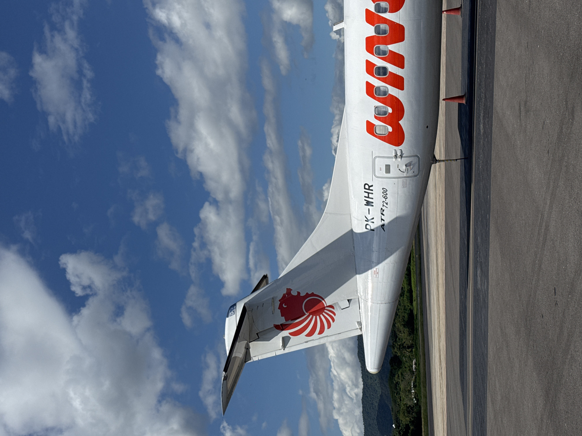 The image shows the tail section of a Wings Air ATR 72-600 aircraft, registration PK-WHR, parked on an asphalt runway at an airport. The plane's tail features the Wings Air logo - a stylized red lion head with radiating stripes. The main body of the plane is painted white with "Wings" in large red letters near the passenger windows. There are orange traffic cones visible next to the plane. The sky is blue with scattered clouds in the background. A strip of green vegetation can be seen in the distance, indicating the airport is near a forested area. The presence of Wings Air, the ATR 72-600 aircraft model, and the runway setting suggest that the location may be in Labuan Bajo, Indonesia, as Wings Air operates flights to and from the Komodo Airport in Labuan Bajo.