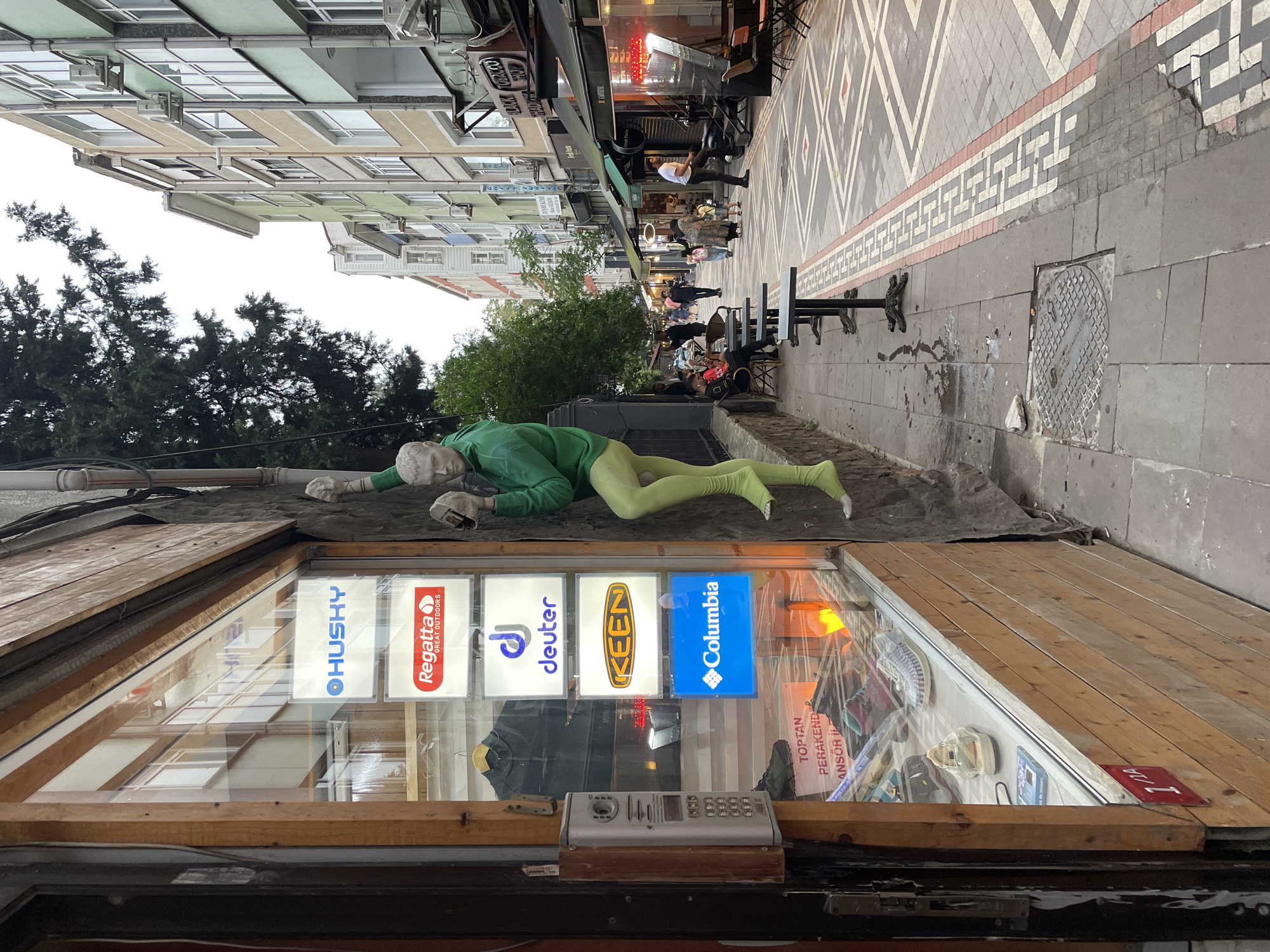 The image shows a street in Kadıköy, Turkey, featuring a mannequin dressed in a green outfit, appearing to climb a textured wall. The mannequin is positioned in front of a shop window displaying various branded items, including signs for Husky, Regatta, Deuter, Keen, and Columbia. Inside the shop, shoes and clothing are visible. The street itself is paved with decorative tiles. Several people are walking along the sidewalk, and tables and chairs are set up outside a business. Buildings line the opposite side of the street, with awnings and signage indicating shops. The sky is overcast.