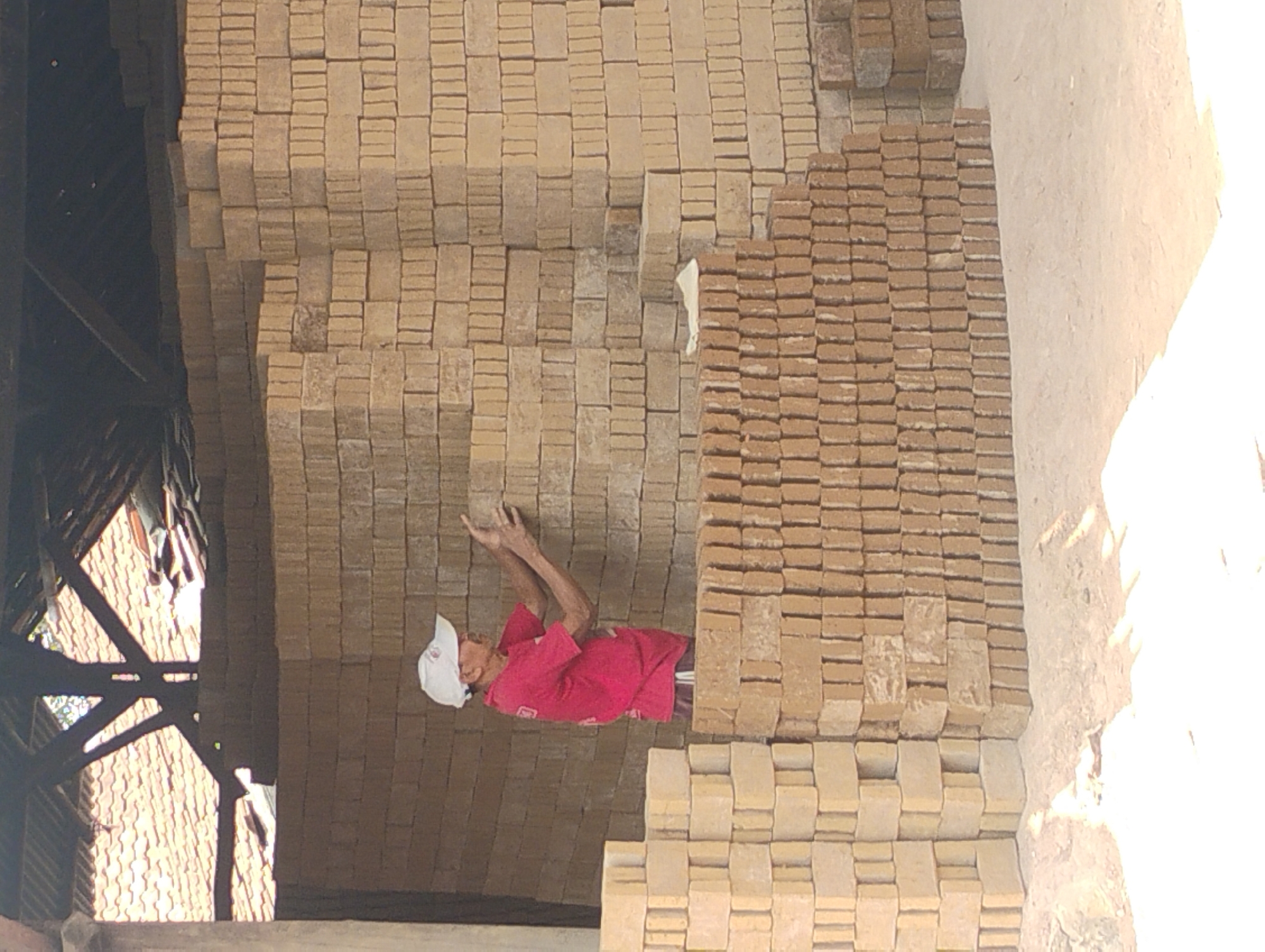 An elderly man wearing a white cap and a red t-shirt is stacking yellow bricks under a rustic, open-sided structure with a corrugated tin roof supported by wooden beams. He is positioned among tall stacks of bricks. To the left of the man, there are stacks of bricks reaching approximately waist height, and to his right, the stacks are significantly taller, obscuring him from view from the chest up. The man's hands are extended, appearing to adjust a brick. The environment is dry and dusty, with visible sand and dirt on the ground. The overall scene suggests a brick-making or storage area.