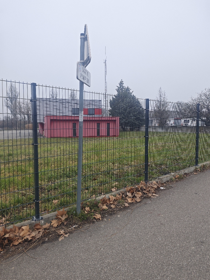 The image depicts an exterior scene in Hoenheim, France, under an overcast sky. In the foreground, an asphalt path or sidewalk is visible on the right, bordered by a strip of ground containing sparse green vegetation and numerous fallen brown leaves, particularly concentrated along the base of a black metal fence. This fence, composed of vertical bars and horizontal supports, extends across the midground, separating the paved area from an open grassy field.
A grey metallic pole is positioned in front of the fence, slightly to the left of the frame's center. Attached to this pole is a rectangular white sign with black text, oriented vertically, which reads "SORTIE POMPIERS" (Fire Exit / Firefighters' Exit).
Beyond the fence, the grassy field appears unmaintained, exhibiting variations in green and brown patches. In the background, a building complex is visible. The most prominent structure features a red lower section with multiple dark, rectangular window or panel openings, surmounted by an upper section with horizontal grey or silver siding and additional dark windows. To the right of this building, a large, dark green coniferous tree stands. Further in the distance, a tall, slender communication or transmission tower with a lattice structure is discernible against the light grey sky. Bare deciduous trees are also visible on both the left and right sides of the background.
No individuals, actions, or direct interactions are present in the scene. The overall lighting is diffuse due to the overcast conditions.