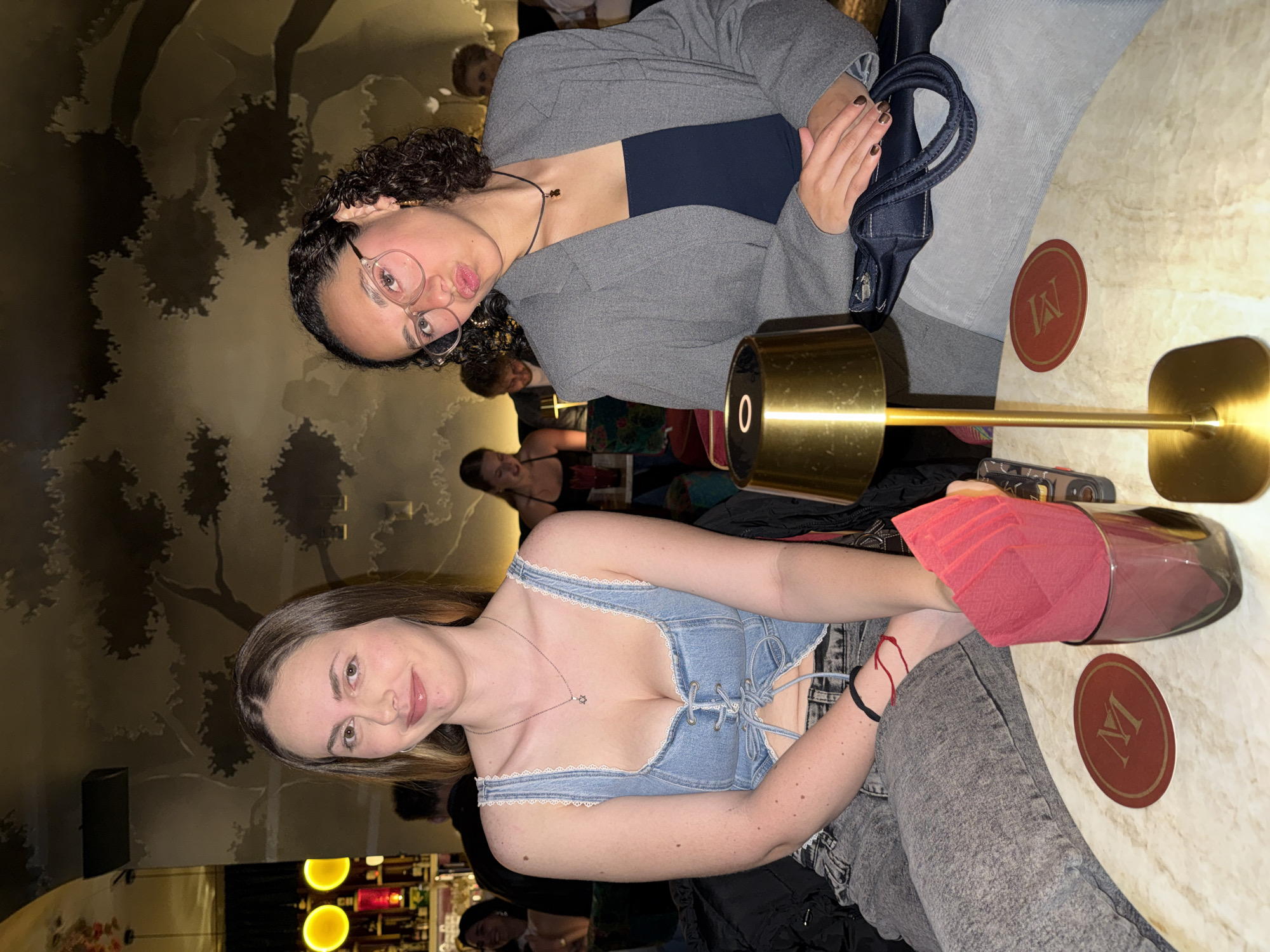 Two women are seated at a marble-topped table in a restaurant in Mexico City, Mexico. One woman is wearing a blue denim top with white lace trim and grey jeans, and has a red bracelet on her left wrist. A pink napkin is tucked inside a glass with a stainless-steel base next to her. The other woman is wearing a grey blazer over a navy top, with grey jeans, and is holding a dark blue purse. A gold lamp is positioned between them. The table has a circular coaster with a stylized "M" on it. In the background are other patrons, a wallpaper mural of trees, and a bar with bottles.