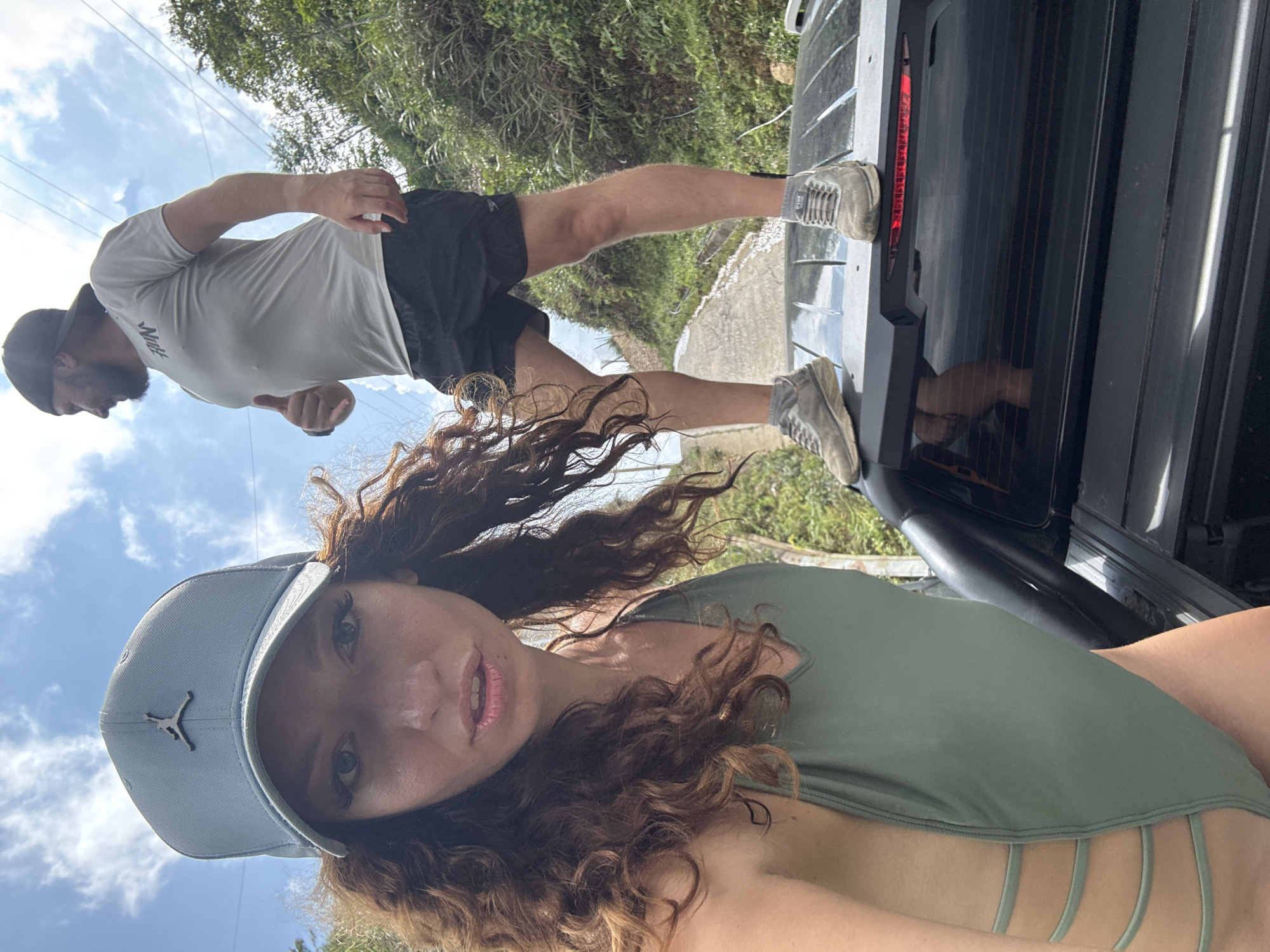 The image depicts a woman wearing a light-colored Jordan cap and a green swimsuit, likely a selfie, with a man standing on the open tailgate of a black SUV in the background. The man is wearing a light grey short-sleeved shirt and black shorts, and grey sneakers. He is standing on the tailgate with his back facing the camera. The surrounding environment appears to be a lush, green, possibly mountainous area. Power lines are also visible, and a glimpse of a paved road is visible behind the vehicle. Given the context, this is likely in a rural area of Suối Giàng, Vietnam.