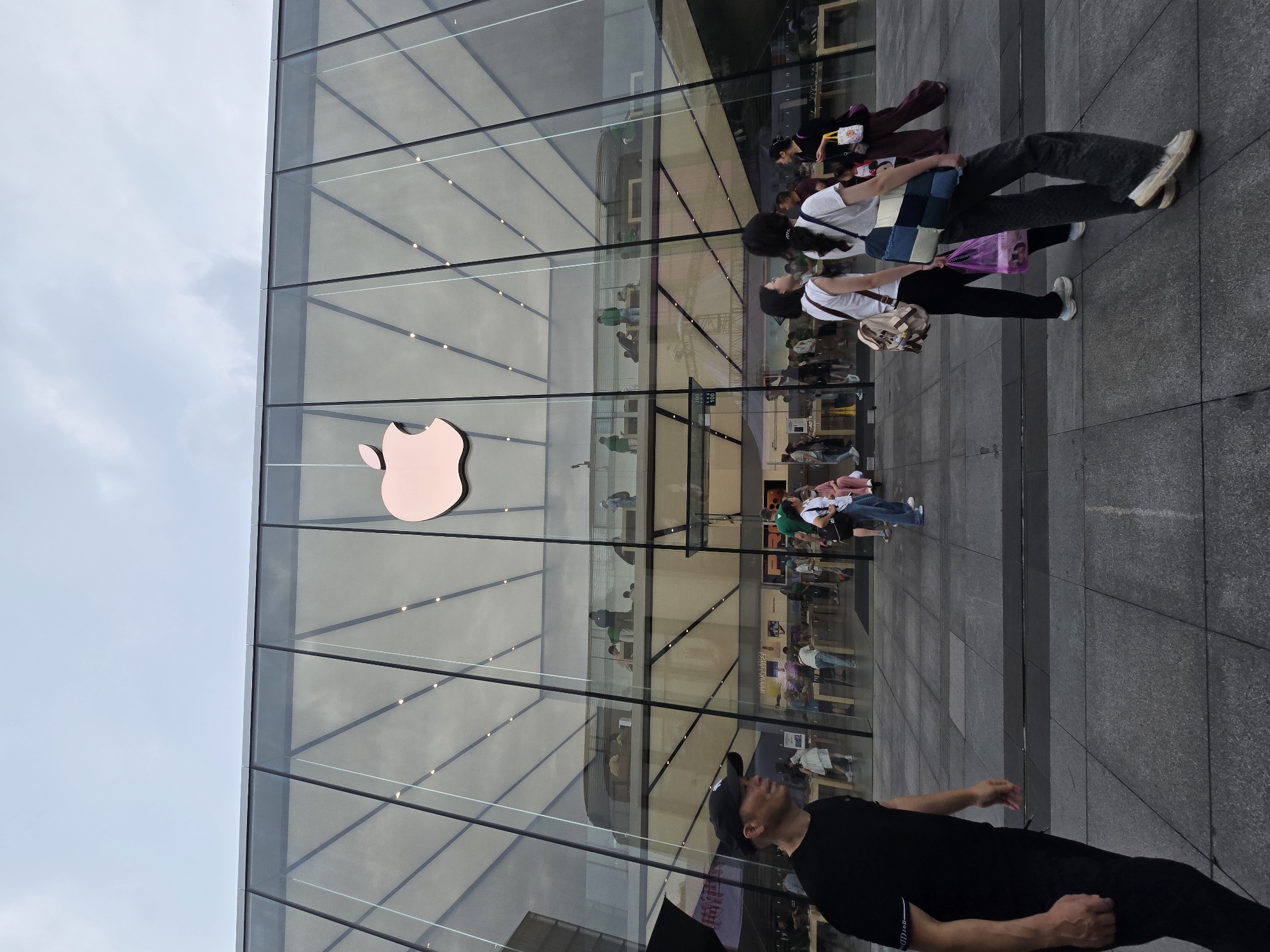 The image captures the exterior of a modern, multi-story Apple Store in an urban setting, featuring a prominent glass facade. A rose gold Apple logo is affixed to the left side of the building's glass exterior. The ground in front of the store is paved with dark grey tiles, indicating a pedestrian area. The sky above is a light, overcast grey, suggesting daytime with diffuse lighting.
Several individuals are present outside the store. In the bottom right foreground, a man wearing a black t-shirt and a black baseball cap is looking upwards towards the building. To his left, a group of three people walks from left to right across the frame. One person wears a white t-shirt, black pants, and carries a beige shoulder bag. Another, slightly ahead, wears a white t-shirt, light grey wide-legged pants, and carries both a patchwork denim-style bag and a translucent purple shopping bag. A third person, partially visible behind them, wears a dark outfit and carries a white shopping bag.
The transparent glass walls offer views into the store's interior, revealing multiple levels of retail space with display tables and numerous customers and staff. Reflections of the surroundings, along with the bustling activity inside, are visible across the glass panels. Some staff members appear to be wearing green shirts. The scene depicts a lively commercial environment in Shangcheng District, China.