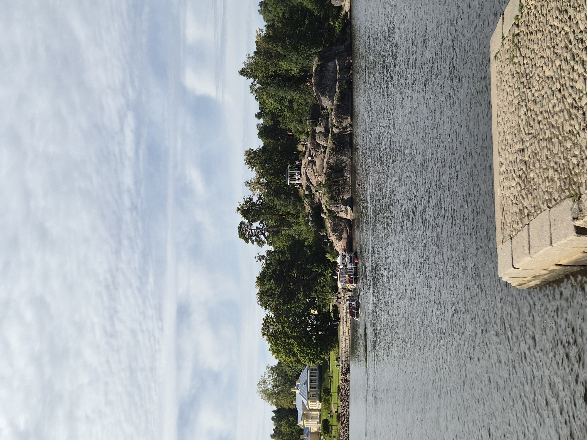 The image shows a scenic view of the Vyborg Bay in Vyborg, Russia. In the foreground, a cobblestone embankment juts into the dark, rippling water. Across the bay, a rocky island is covered with dense green trees and a white gazebo. Several people are gathered on the island, some standing near the gazebo and others scattered on the rocks. A light yellow building with white columns and a gray roof is visible on the shore to the left, bordered by a manicured lawn and trees. Two small boats are docked near the shore of the island, with people on board or disembarking. The sky above is a mix of white clouds and blue. The overall impression is a tranquil summer day in a park-like setting by the water.