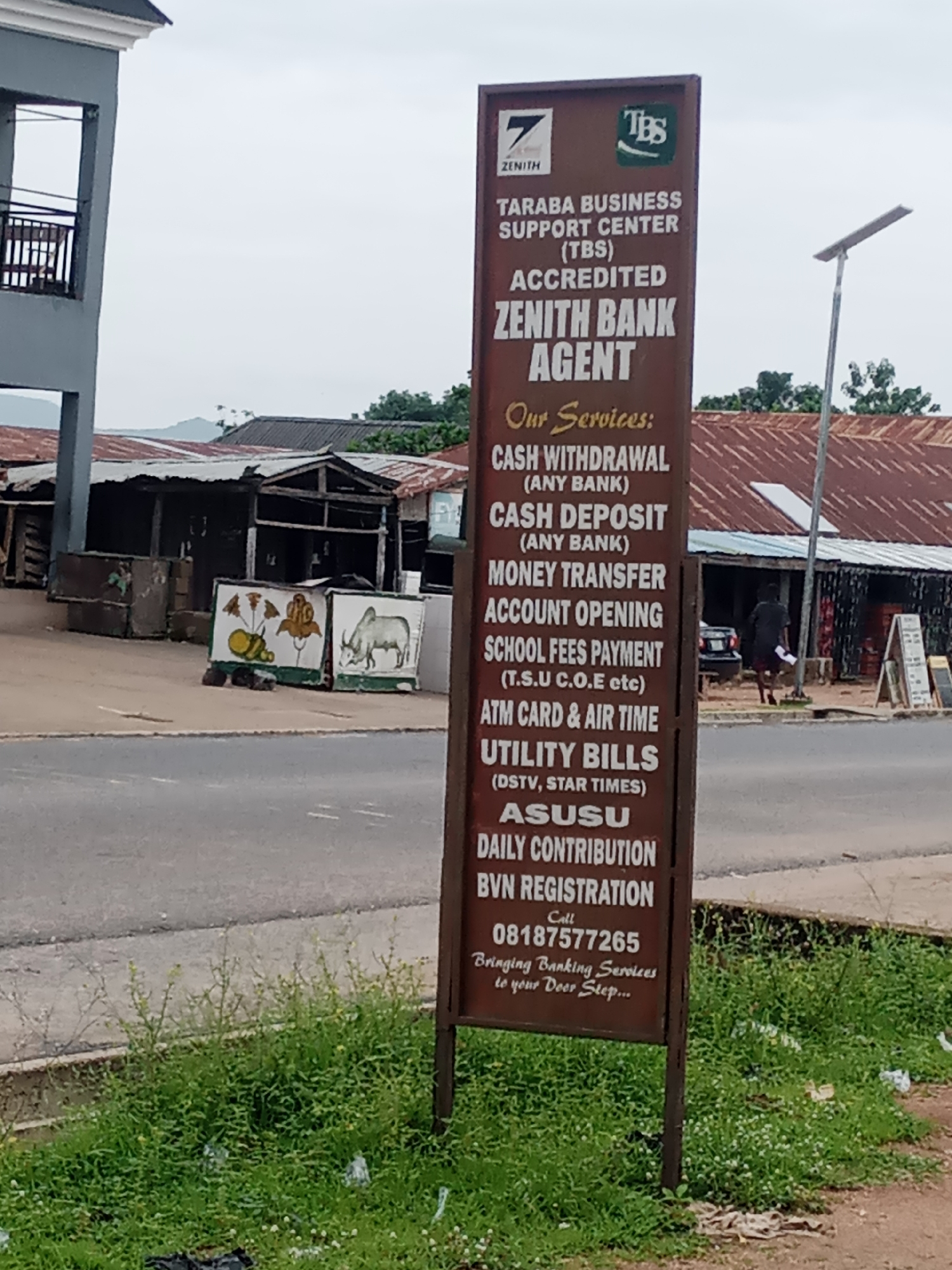 The image captures a Zenith Bank agent sign located in Jalingo, Nigeria. The sign, predominantly brown with white lettering, lists various financial services offered, including cash withdrawal and deposit, money transfer, account opening, school fees payment, ATM card and airtime purchase, utility bill payment, ASUSU daily contribution, and BVN registration. A phone number, "08187577265," is prominently displayed, along with the tagline "Bringing Banking Services to your Door Step..." The sign also features logos for Zenith Bank and "TBS" (Taraba Business Support Center).
In the background, there are modest, zinc-roofed structures, typical of a local business area. A street with a paved surface is visible in the foreground. A solar-powered street light stands to the right of the sign. A person, casually dressed, is walking in the middle distance towards the right side of the frame, carrying what appears to be a piece of paper. The environment appears to be outdoors, with some green vegetation and scattered debris in the foreground. The sky is overcast.