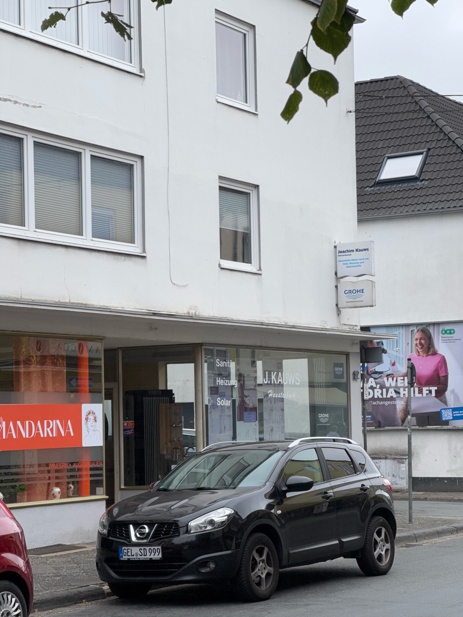 The image shows a street scene in Cleves, Germany. In the foreground, a black Nissan Qashqai is parked at the curb, its license plate reading "GEL SD 999". To the left, the front of a red car is visible. Behind the parked car is a white building with storefronts at ground level. One storefront has a large orange sign with the word "MANDARINA" in white lettering. Next to it, another storefront displays signs for "Sanitä", "Heizung", and "Solar" along with the "J. KAUWS Haustechnik" logo and "GROHE WASSERTECHNOLOGIE". Further to the right on the building, two white signs are mounted, one reading "Joachim Kauws Meisterbetrieb Innovative Ideen rund um Sanitär, Heizung und Solartechnik" and below it another "GROHE WASSERTECHNOLOGIE" sign. A large billboard advertising "JA, WEILORIA HILFT" is visible on the right side of the building. The street is paved, and a sidewalk is present. The sky appears overcast. The building itself has multiple stories with windows.