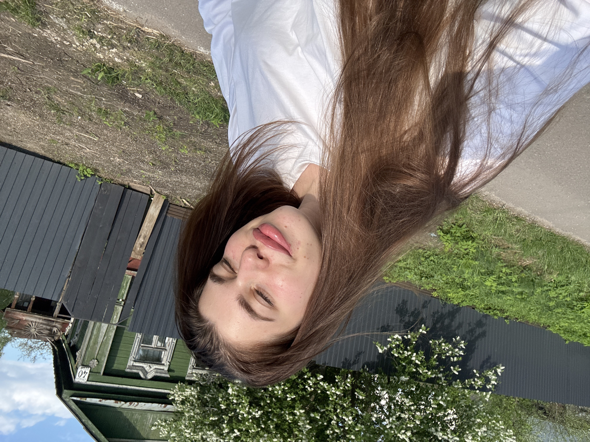 A young woman lies down in an outdoor setting in Мещёры, Russia. Her long brown hair is spread out around her. She wears a white shirt. Her eyes are closed. To the left of the woman is a dark gray wooden fence and a green wooden house with white trim. The number "40" is visible on the house. To the right, a flowering tree is near the woman. Beyond the woman's head is a gray concrete walkway. Beyond that is ground covered with dirt and vegetation.