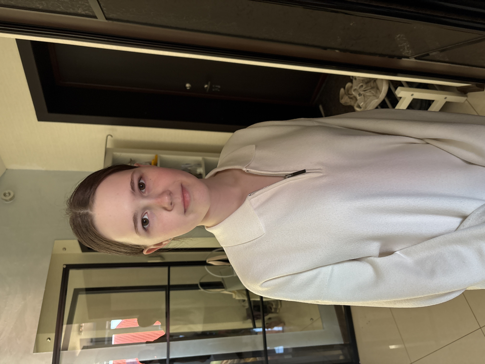 A young person with light skin and brown hair, parted in the middle, is wearing a white zip-up sweater. They are looking at the camera. The background includes a black door frame, a glass-paneled structure, and a light-colored wall. The location appears to be indoors, with tiles visible on the floor. Given the context of Ulyanovsk, Russia, it may be assumed that the setting is a domestic or public interior within the city, but nothing further is confirmed about that.