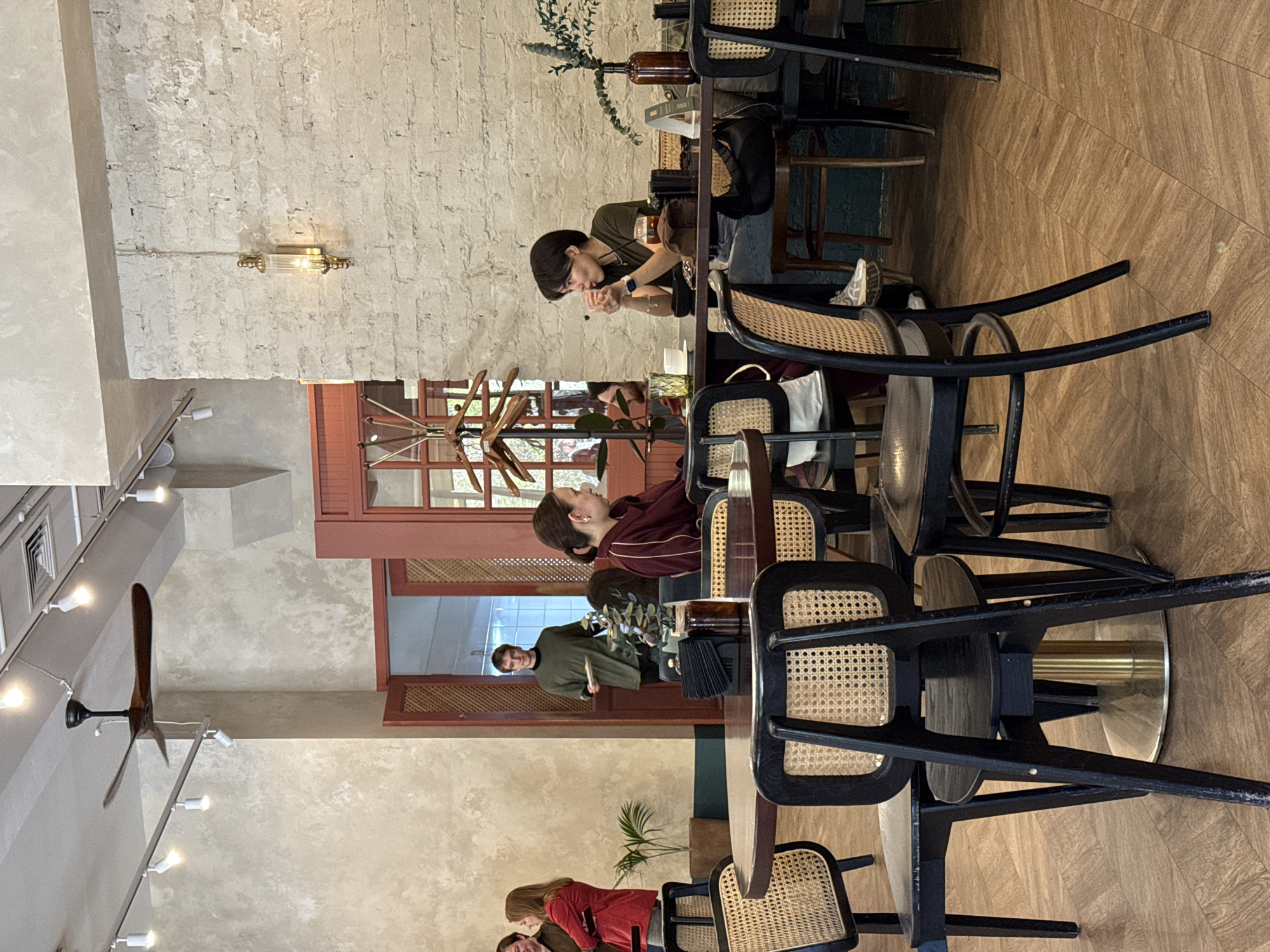 The photograph depicts an interior scene of a café or restaurant, likely in Krasnoyarsk, Russia. Several individuals are visible, seated at tables. At the table closest to the foreground, a person is wearing a red top, interacting with another individual next to them. At the mid-ground table, a woman in a red and white striped top is seated. A person in a green shirt can be seen through the interior doorway. The café features exposed brick walls and vintage-style lighting. The furniture consists of tables and chairs with a distinctive woven cane design. A wooden ceiling fan is mounted on the ceiling. The flooring appears to be wooden parquet.