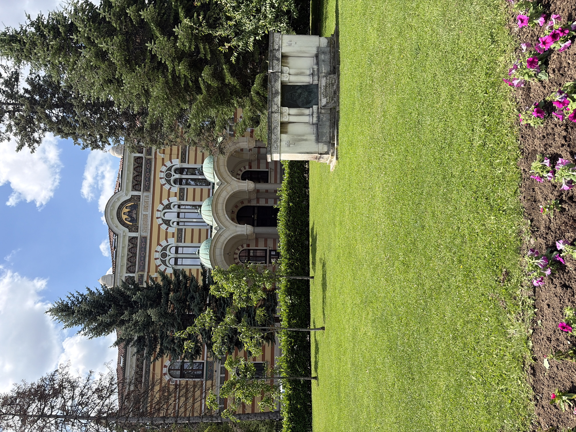 The image shows the building of the National Palace of Culture in Sofia, Bulgaria, on a sunny day with a blue sky and white clouds. The building is yellow with ornate details and a red tiled roof. In front of the building is a well-maintained lawn with several young trees. To the right of the lawn is a stone monument with a bronze plaque. In the foreground, there is a flower bed with pink and purple petunias. The scene depicts a serene and beautiful public space.