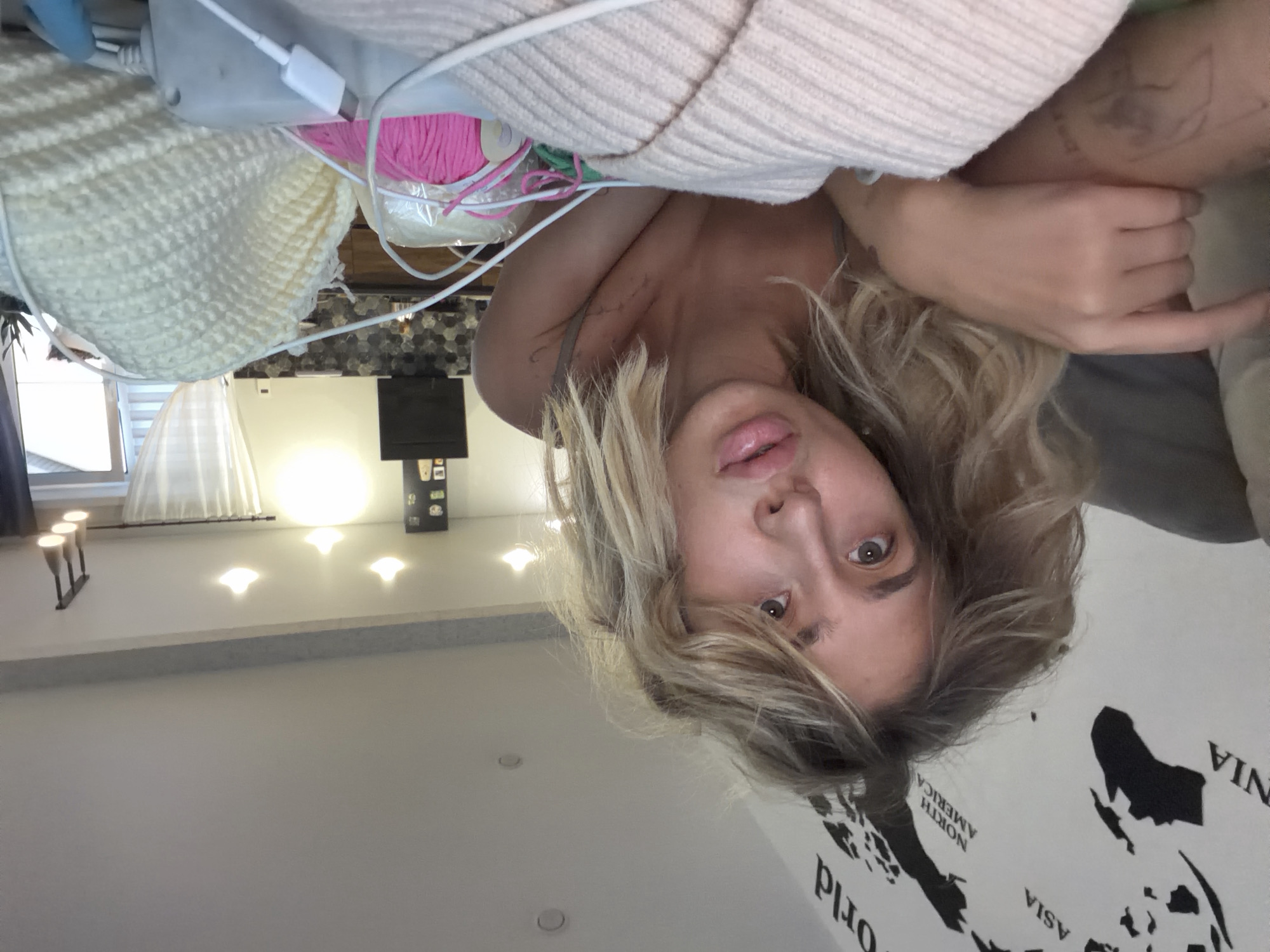 A young woman with blonde, wavy hair is lying down in what appears to be a living room. She is looking directly at the camera with her mouth slightly open. Her left arm is extended, resting on a light-colored knitted object. Various skeins of yarn in pink and yellow, along with charging cables, are visible in the foreground. In the background, a world map is adhered to the wall. The lighting suggests it is daytime. The scene is consistent with a domestic setting, likely a home in Kamenka, Russia.