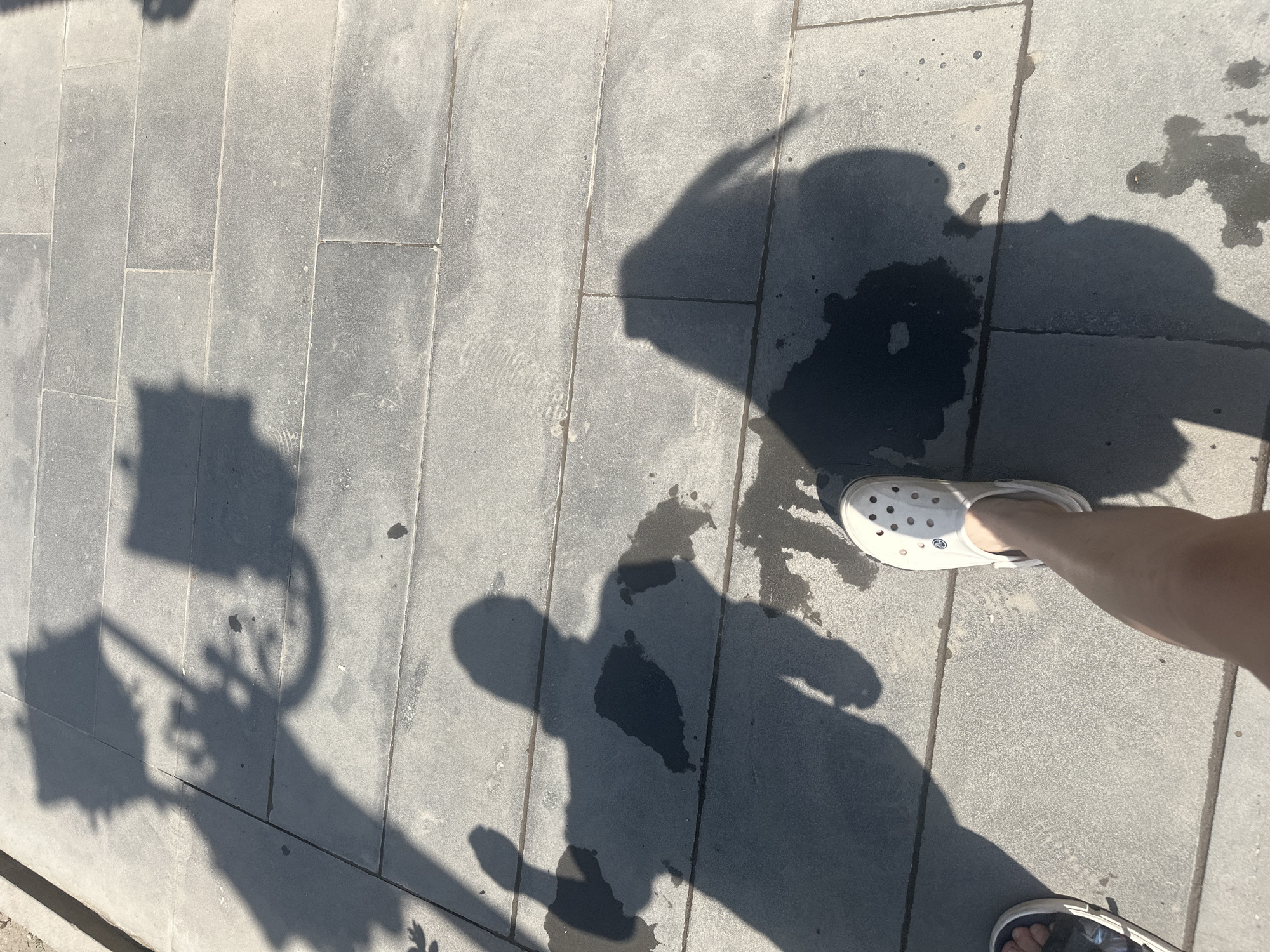 The image depicts a street scene with shadows cast on a grey, tiled pavement. The shadows suggest the presence of a street lamp or similar structure in the background, as well as two individuals. One individual's shadow is clearly visible, showing a person with an arm raised. The feet of two individuals are also visible in the foreground, one wearing a white Croc and the other a sandal. There are dark patches on the pavement, possibly indicating wet spots from recent rain or spilled liquid. The lighting suggests it is daytime, with the sun casting long shadows. This scene could be anywhere with paved streets and sunlight, and there is no specific information linking it to Gyumri, Armenia.