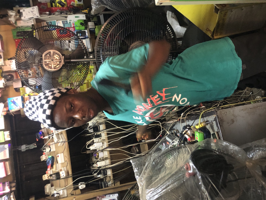 The image depicts an indoor scene in a cluttered electronics shop or charging station located in Enugwu-Ukwu, Nigeria. The central figure is a young adult, appearing female, with dark skin and a noticeable sheen on her forehead and face. She wears a black and white checkered beanie and a teal t-shirt with red and white text partially visible as "LIKE MONEY NO." Her right arm is blurred, indicating motion, with her hand appearing to manipulate a cable or connector.
Behind the individual, a wooden shelving unit occupies the left background, densely packed with various electrical outlets, multi-plugs, power adapters, and packaged electronic accessories. Numerous white and black cables hang vertically from these shelves, creating a tangled network. To the right of the central figure, a large black standing fan with a circular metal grill is prominently positioned, and another partial fan is visible further right. The right background also features shelves displaying packaged electronics.
In the foreground to the left, a counter or workbench holds items including a fan covered in clear plastic, a green object (possibly a multimeter), a yellow-handled screwdriver, and additional cables. The overall environment is functional, characterized by numerous electronic components and wiring, suggesting services related to charging or selling electronics.