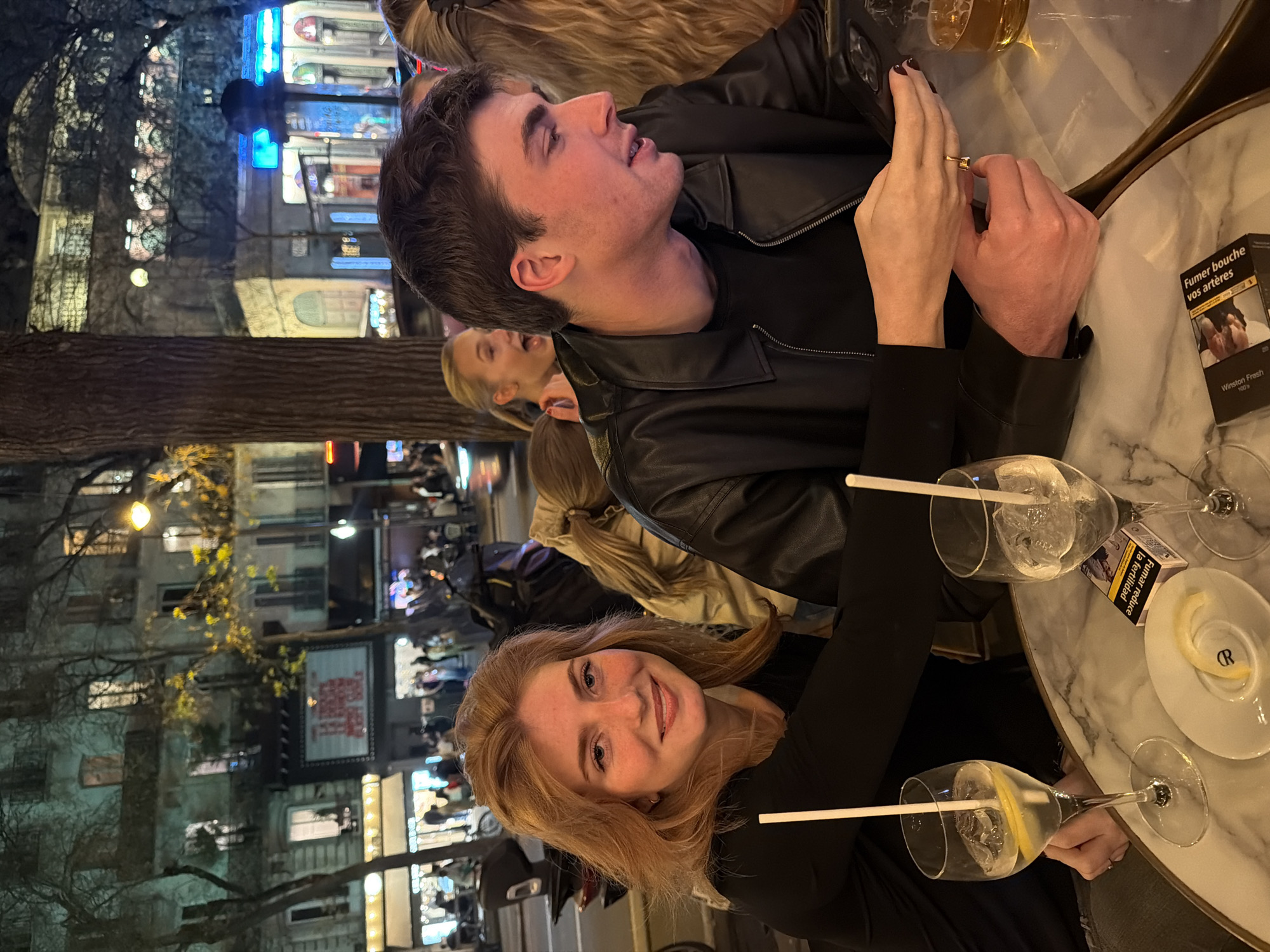 The image shows two people, a man and a woman, sitting at a table outside a restaurant or bar in Paris, France. The woman has reddish hair and is smiling at the camera. She is wearing a black shirt. The man has dark hair and is wearing a black leather jacket. They appear to be on a date. There are two glasses of what appear to be gin and tonics on the table, along with a saucer with a lemon peel and a pack of cigarettes. The table is round with a marble top. In the background, other people are sitting at tables and walking on the sidewalk. Buildings line the street, lit with streetlights and illuminated signs. A large tree trunk is visible in the foreground.