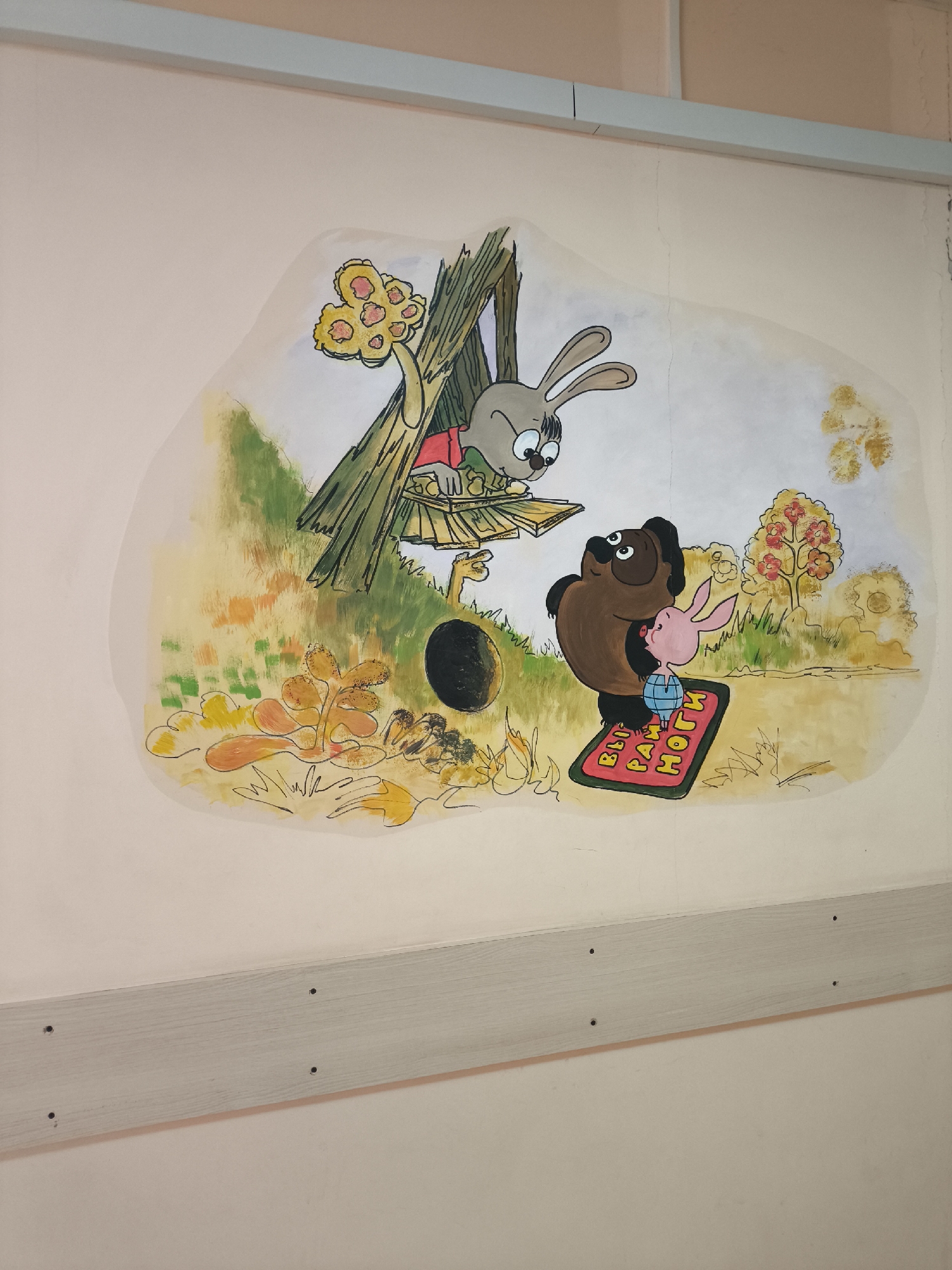 The image features a colorful wall mural, likely hand-painted, depicting characters from the classic Soviet animated series "Vinni-Pukh" (Winnie-the-Pooh). The mural is set against a light-colored wall, with light gray wooden trim running horizontally at the top and bottom.
The scene portrays an outdoor, whimsical environment. In the upper left, a gray rabbit, resembling the "Rabbit" character from the series, peers down from a rustic wooden structure, possibly a treehouse or a collection of logs. It appears to be holding and possibly dropping or handing down flat wooden pieces. On the ground below, a brown bear, akin to "Vinni-Pukh," stands with a small pink piglet, similar to "Pyatachok," next to him. Both the bear and the piglet are looking up at the rabbit.
Near the base of the wooden structure, a dark, circular opening is visible on the ground, suggesting a burrow or den. The background is filled with stylized foliage, patches of green and yellow ground cover, and trees with autumn-colored (yellow and orange) leaves.
A notable object is a red doormat placed on the ground in front of the bear and piglet. Yellow Cyrillic text is written on it, reading "ВЫ РАЗУВАЙТЕ НОГИ," which translates to "TAKE OFF YOUR SHOES" or "UNSHOE YOUR FEET." This indicates the mat serves as a welcoming sign at an entrance.
The mural's style and characters, combined with the visible Russian text, strongly suggest a location in Russia, such as a children's hospital ward, kindergarten, or school in Khimki. The painting shows some signs of age, including minor cracks on the wall surface.