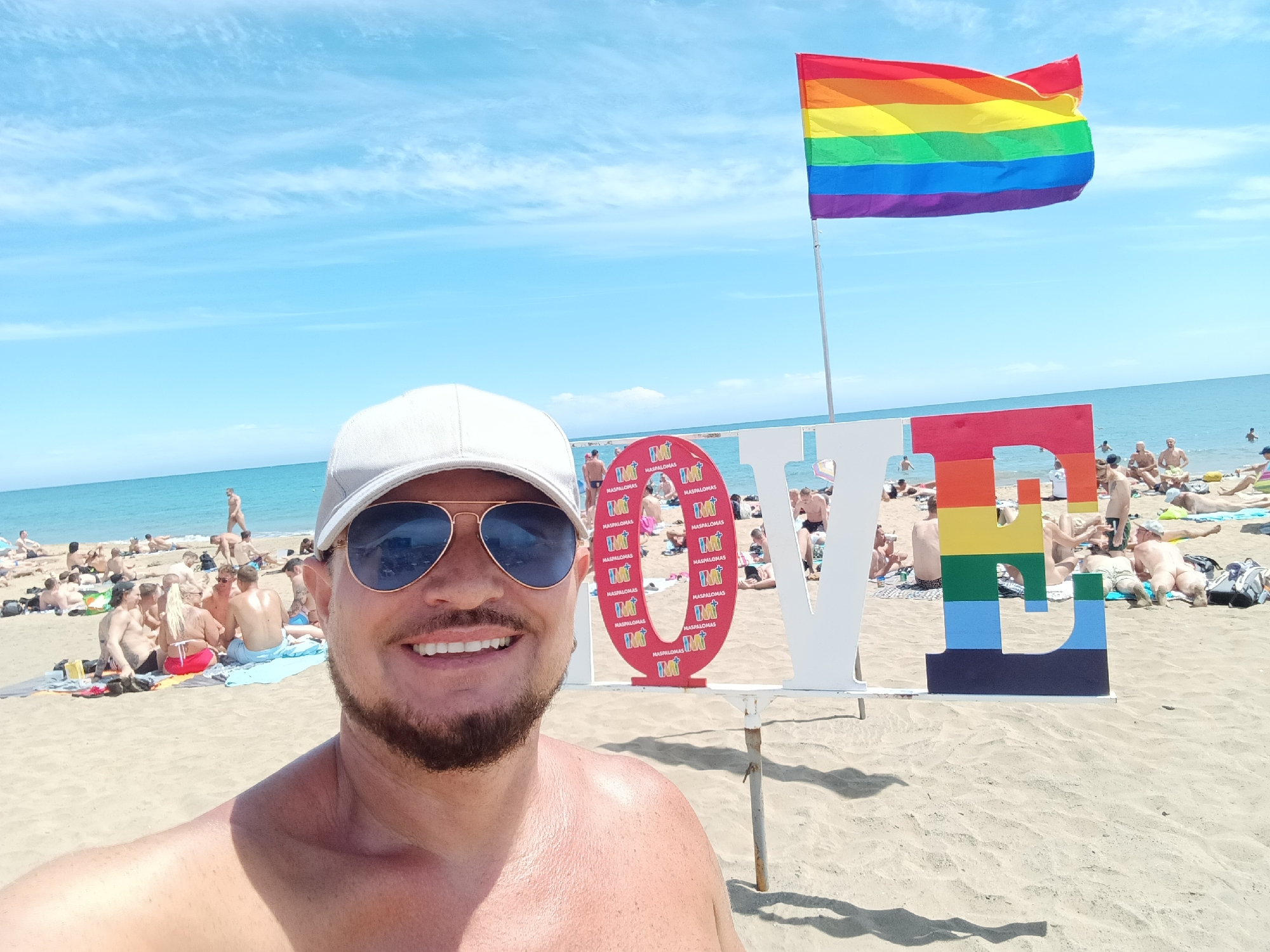 The image is a selfie taken on the beach in San Bartolomé de Tirajana, Spain. A man wearing a white baseball cap and sunglasses smiles at the camera. Behind him is a white "LOVE" sign, with the "O" filled with images of "IMT MASPALOMAS", and the "E" painted in rainbow colors. A rainbow flag flies above the sign. The beach is crowded with sunbathers. The sky is blue with some clouds, and the sea is visible in the background.
