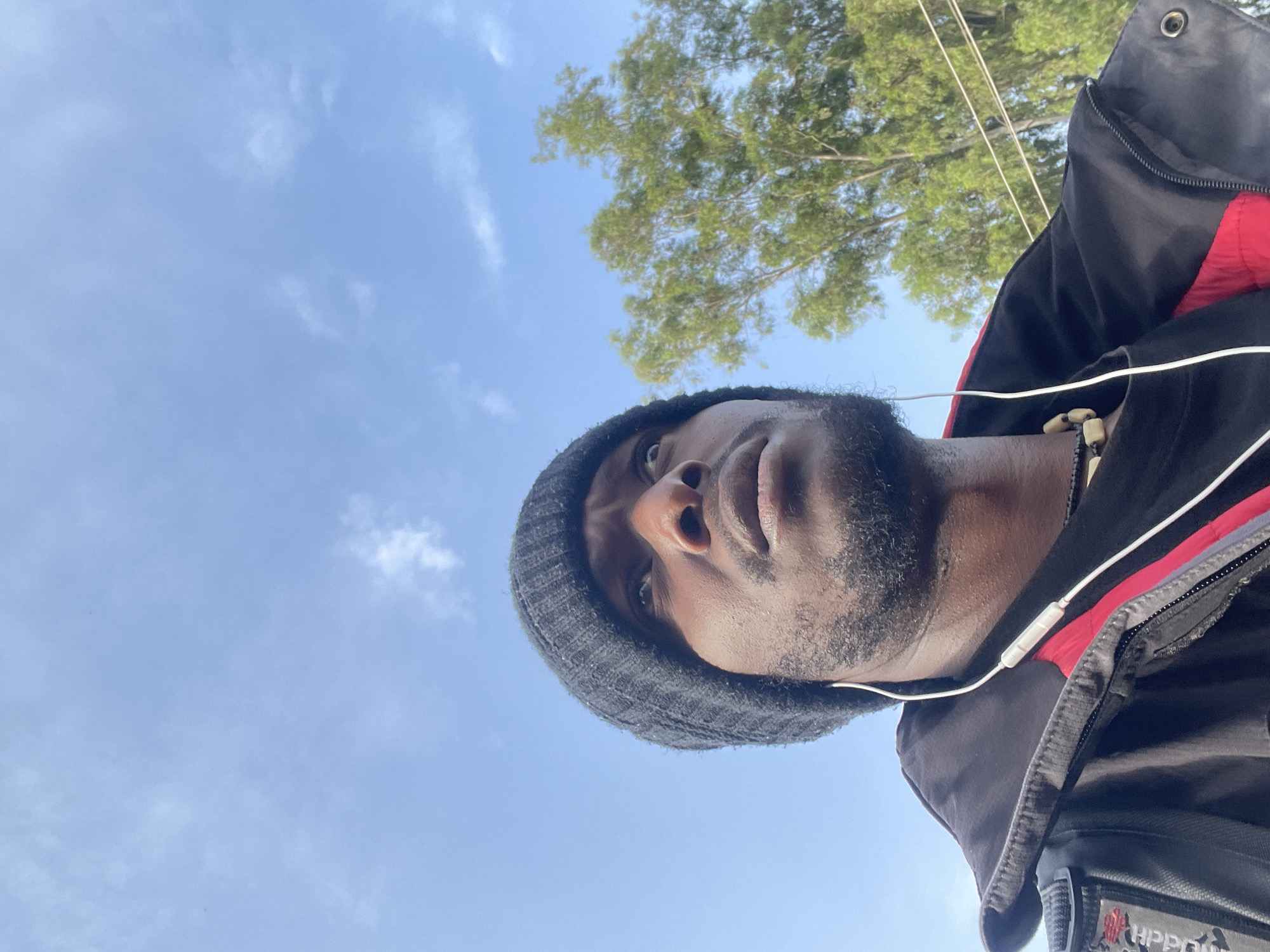 In this close-up, low-angle shot, a Black man wearing a black beanie and a black jacket with red lining looks upwards towards a clear blue sky with scattered white clouds. He has earbuds in his ears and a beaded necklace around his neck. In the background, a green tree with full foliage is visible, along with power lines. The image appears to have been taken outdoors in a location that could be Jos, Nigeria, given the presence of lush greenery and clear skies, typical of the region. The lighting suggests it is daytime, likely morning or afternoon.