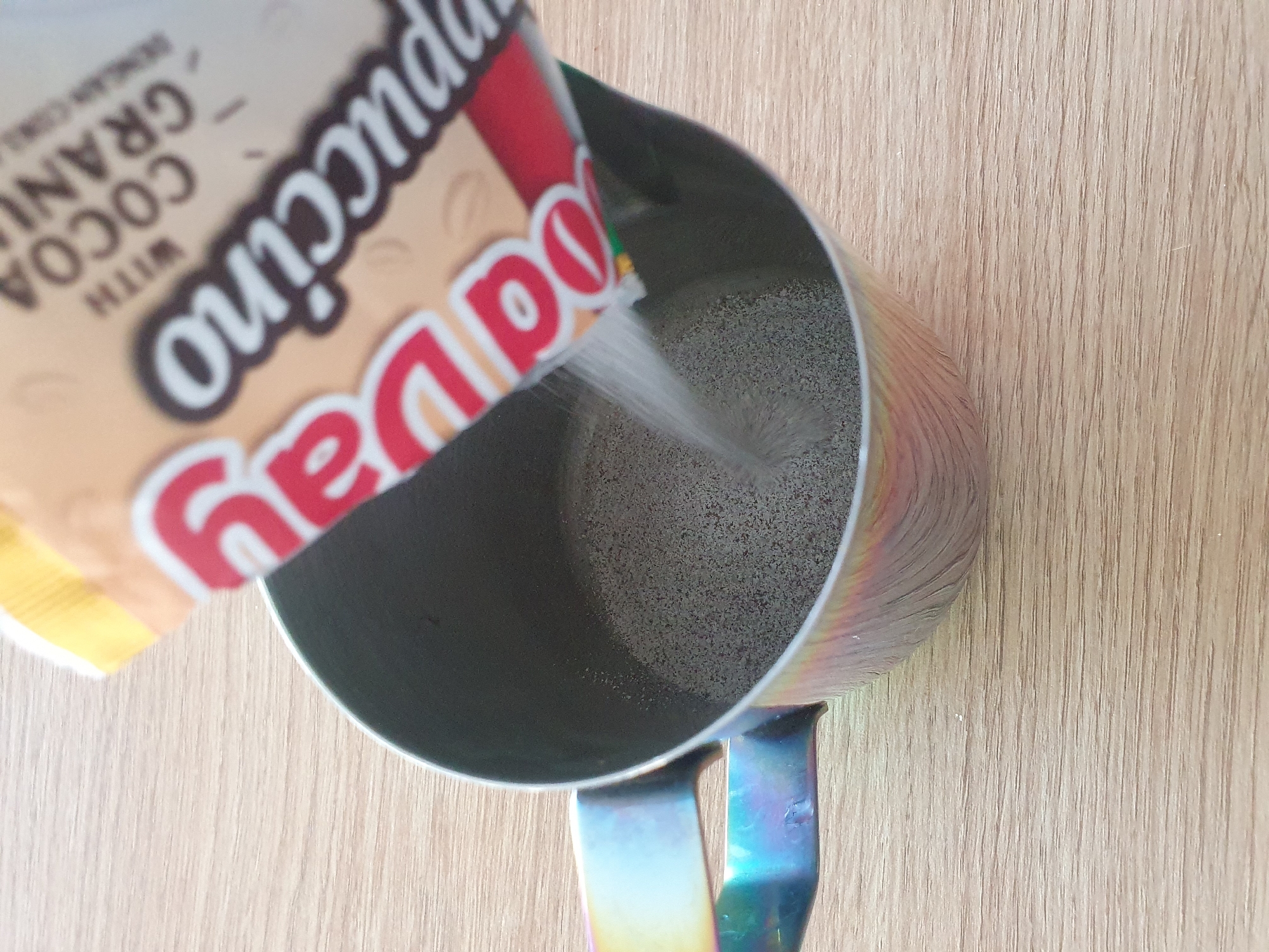 A pouch of instant cappuccino mix is being poured into a metallic pitcher on a wooden surface. The pouch, partially visible at the top of the frame, has red and beige lettering that reads "Cappuccino" and "Cocoa Gran" and "with". The pitcher is cylindrical with a handle that displays a rainbow of colors. The contents of the pitcher appear to be a dry, granular mixture. The lighting suggests an indoor setting, possibly a kitchen.