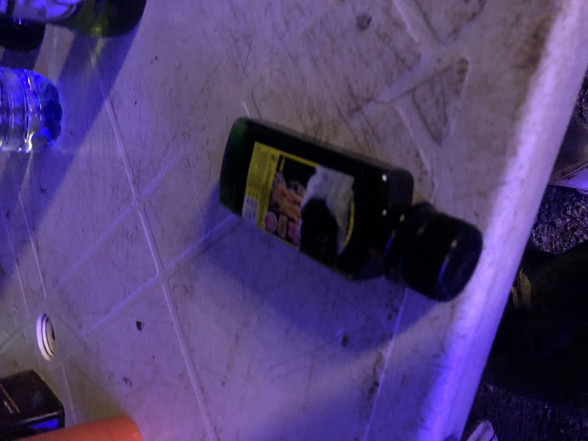The image shows a dark, blurry scene illuminated by purple and blue lights. A dark bottle with a green cap and a yellow label is lying on its side on a textured, off-white surface. The label features images of what appears to be food or possibly an advertisement. To the left of the bottle, a portion of an orange cone is visible, and behind it, a dark object, possibly a speaker or piece of equipment, can be seen. In the background, parts of two water bottles are visible, one clear and one green. The ground beneath the table is dark and appears to be covered in gravel or rough material. The overall impression is that of an outdoor gathering or event taking place at night, possibly a party or a festival, given the lighting and the presence of drinks and equipment.