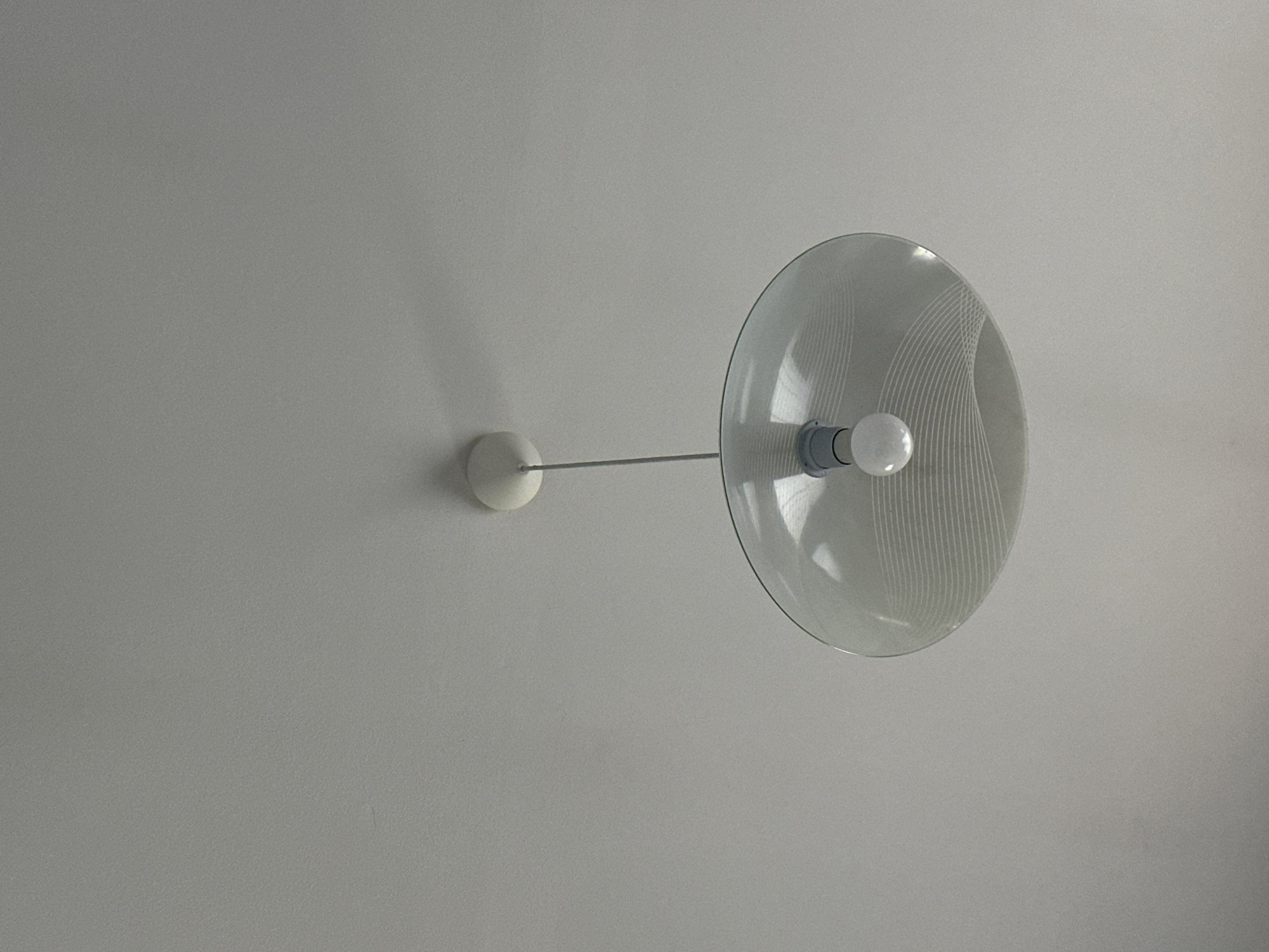 The image shows a close-up of a white ceiling with a single, modern pendant light fixture hanging from it. The fixture has a white circular canopy attached to the ceiling, from which a thin grey cord descends. At the end of the cord is a white socket holding a clear, round light bulb. The light bulb is enclosed by a large, translucent, dish-shaped shade made of glass. The shade has a delicate pattern of white, wavy lines etched or printed onto its surface. The background is the plain, textured white ceiling, with a soft shadow cast by the light fixture on the upper right side. The scene appears to be an interior setting, with no indication of the city of Marten, Bulgaria.