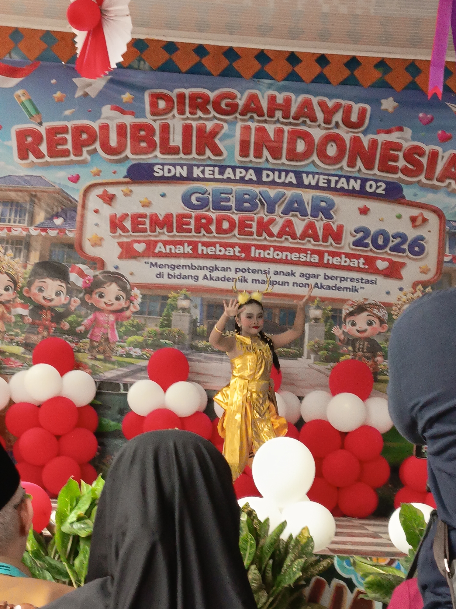 The image depicts a celebratory event, likely a school gathering, with a large banner in the background that reads "DIRGAHAYU REPUBLIK INDONESIA" and "GEBYAR KEMERDEKAAN 2026". The banner is colorful and features cartoon-like illustrations of children in traditional Indonesian attire. Below the main text, there is a smaller inscription that says "Anak hebat, Indonesia hebat." and a quote in Indonesian.
In the foreground, a young girl dressed in a golden, sequined traditional costume is dancing. She has yellow accessories on her head and is striking a pose with her arms raised. Behind her, a group of red and white balloons are arranged in clusters, decorating the stage area. Several people are visible in the foreground, including a person in a black hijab and another partially visible person on the right side of the frame wearing a blue garment. The environment appears to be indoors or a covered outdoor space, given the presence of the banner and the lighting. There is no specific visual information to pinpoint the location within East Jakarta, Indonesia.