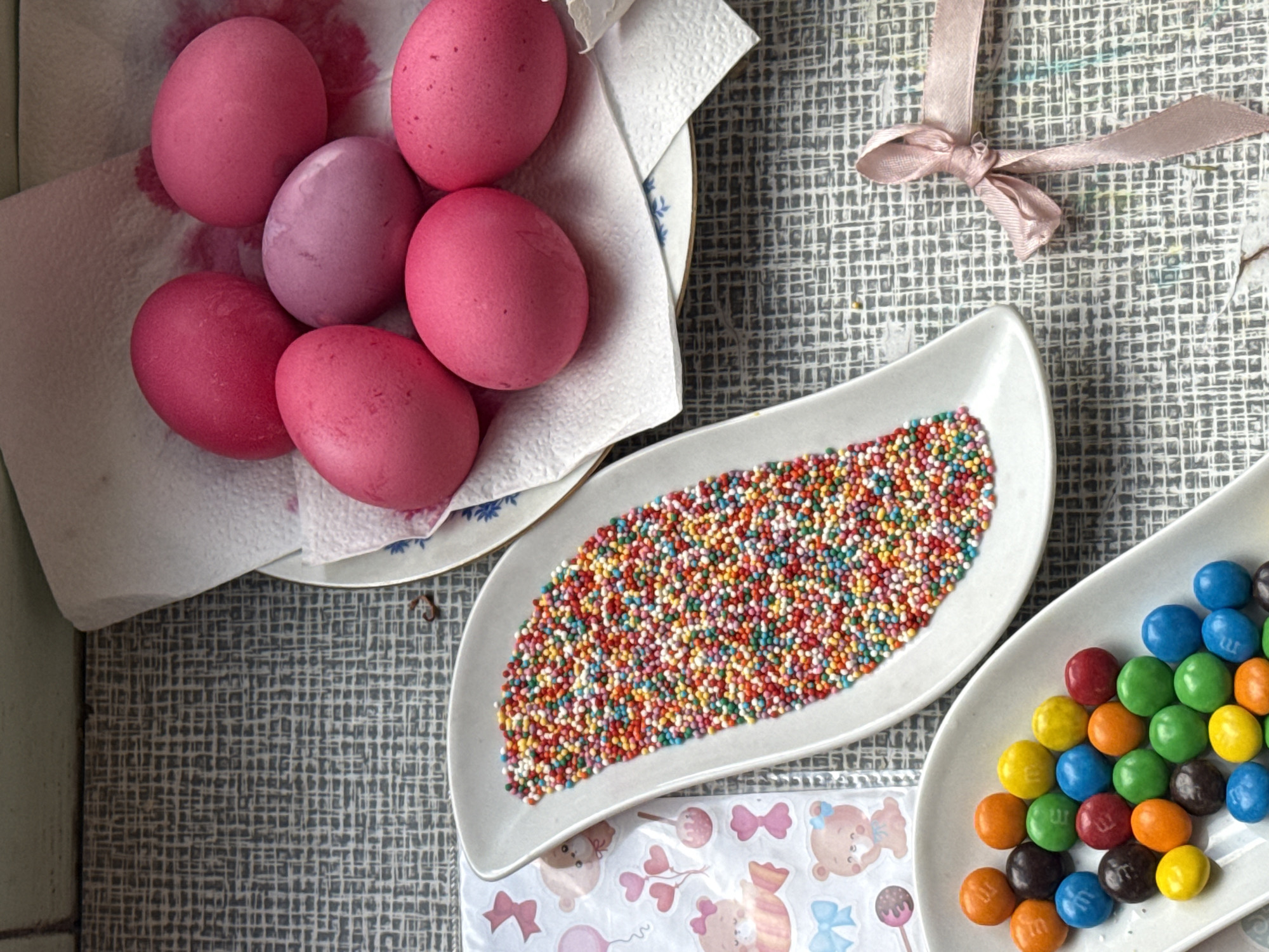 The image shows a table setting with Easter egg decorating supplies. There is a plate with six dyed eggs, mostly pink with one lavender, sitting on a paper towel. There is a white, leaf-shaped dish filled with colorful sprinkles, and another similar dish containing M&M candies in various colors. A pink ribbon with a bow is also visible. The setting includes a patterned gray table cloth. There's a sticker sheet with teddy bears, bows, and candies. The location and city of Yerevan, Armenia are not determinable from the image.