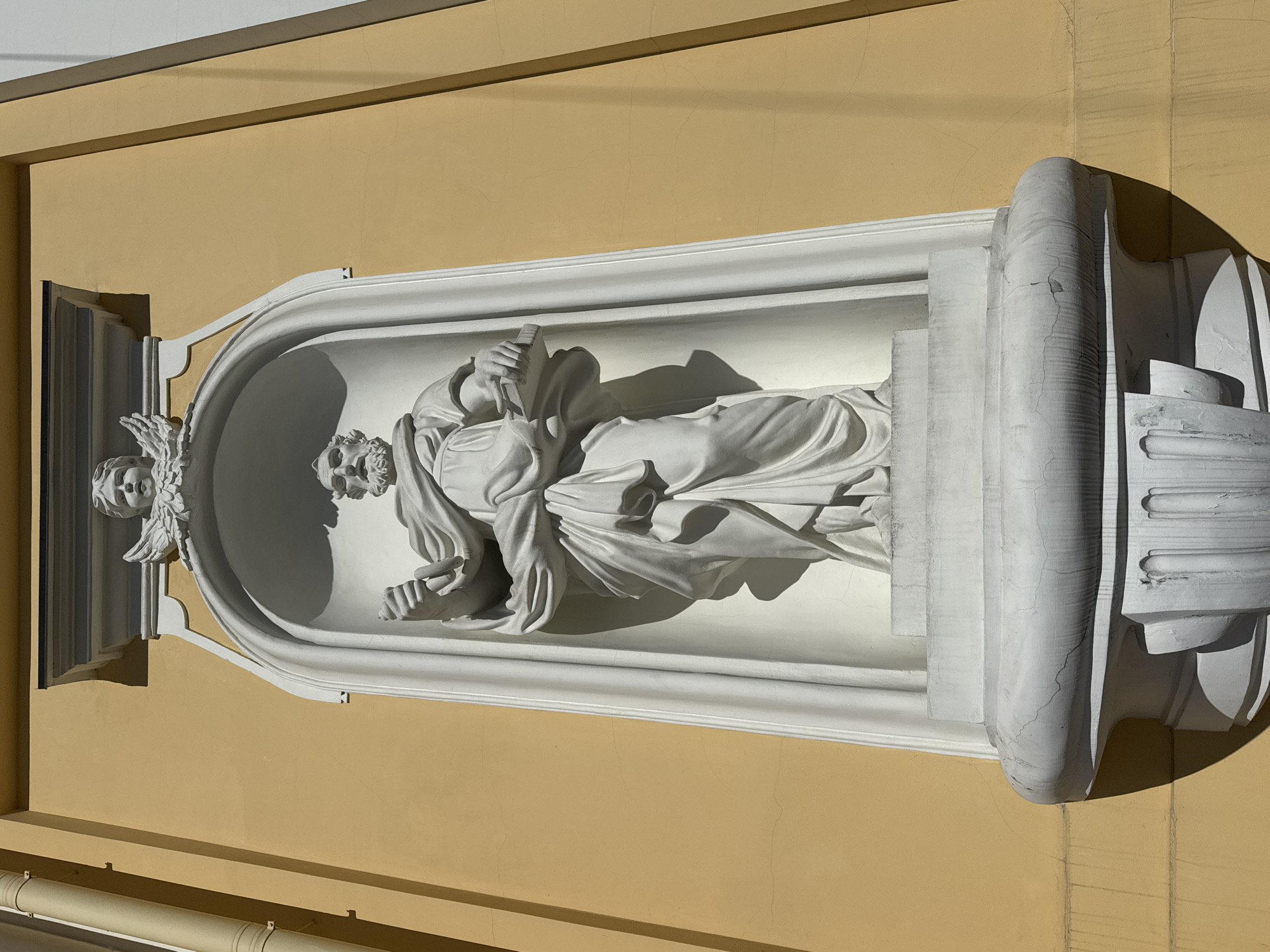 A white sculpted statue of a male figure is prominently displayed within a decorative niche on a building facade in Saint Petersburg, Russia. The figure, possibly a saint or a philosopher, is depicted standing, with a beard and draped in flowing robes. He holds an open book in his right hand, while his left hand is slightly raised. To the left of the main figure's head, a smaller, winged angelic or cherubic head is carved into the niche.
The statue and its surrounding elaborate, arched niche are crafted from white stone or plaster, showing signs of weathering with visible cracks on the surface, particularly on the decorative elements. The niche is recessed into a building wall painted a warm, light yellow or ochre. The wall itself displays faint hairline cracks. Strong, direct sunlight illuminates the scene, casting sharp, distinct shadows to the right and beneath the statue and its architectural framing, indicative of a bright day. No text is visible in the image.