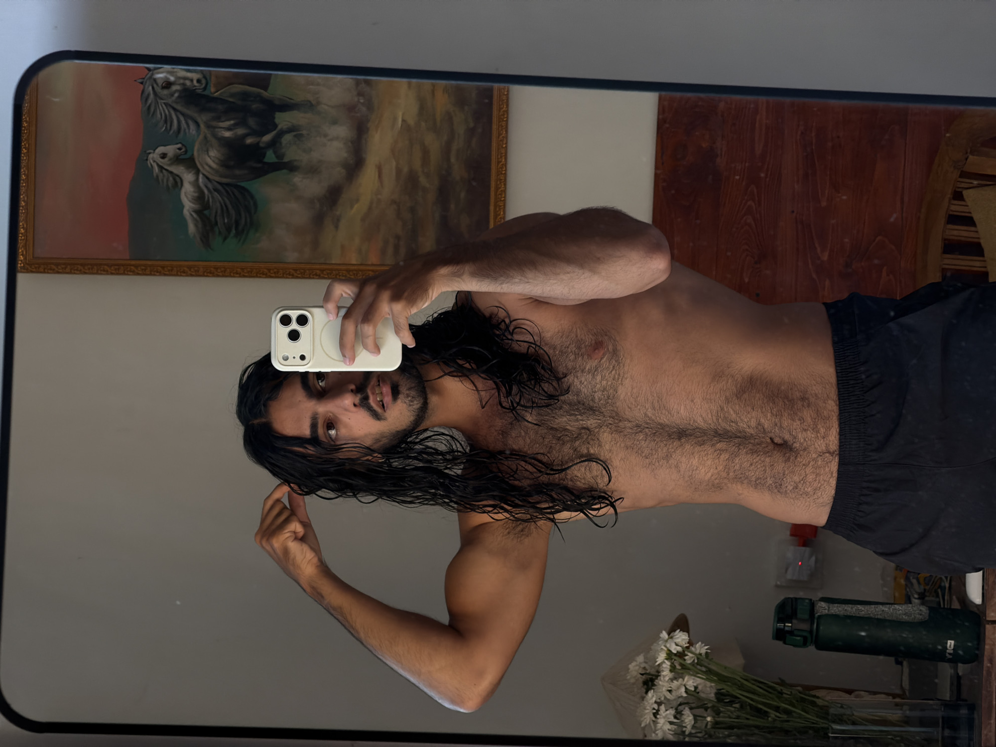 A man is taking a selfie in a mirror in what appears to be an indoor setting in North Kuta, Indonesia. He is shirtless, wearing dark shorts, and has long dark hair. The phone he is using to take the picture has a white case. The reflection in the mirror shows a painting of horses on the wall, wood paneling, flowers in a vase, and a green water bottle, possibly on a table or counter. The wall visible in the mirror is plain.