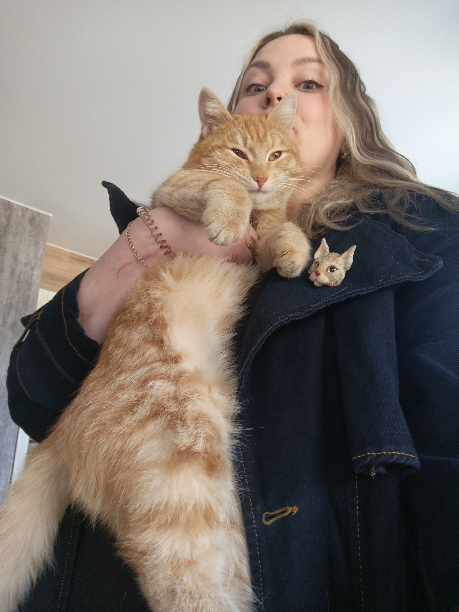 A fair-skinned person with blonde hair is holding a ginger cat. The person is wearing a denim jacket with a cat-shaped brooch on the lapel. The cat appears to be resting its paws on the person's hand. The environment is nondescript. There is no information to support that the image was taken in Noviy, Russia.