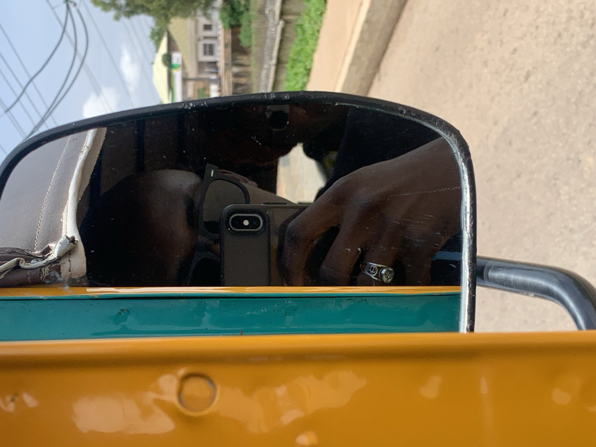 The image is a close-up shot of a person taking a selfie in the side mirror of a vehicle, likely an auto-rickshaw, in Gura Top, Nigeria. The vehicle is predominantly yellow with a turquoise stripe running vertically along its side. The mirror itself reflects the driver, who is wearing dark sunglasses and a ring on their left hand. They are holding a smartphone to take the picture. The background visible in the mirror shows a street scene with buildings, trees, and power lines, indicative of an urban or semi-urban environment. The overall lighting suggests it is daytime.