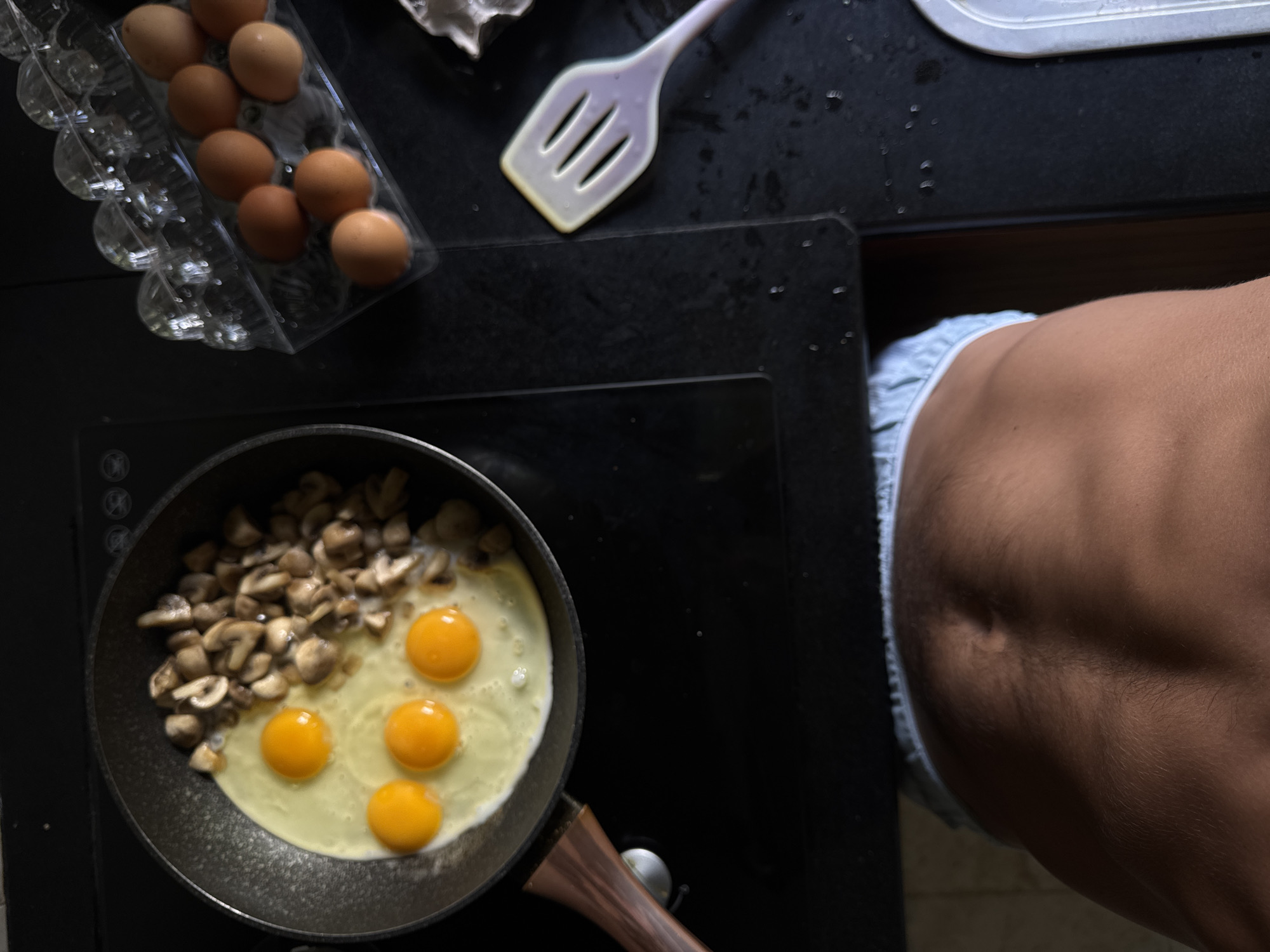 The image depicts a close-up, overhead view of a person preparing breakfast in what appears to be a kitchen. The primary subjects are a frying pan containing three eggs and chopped mushrooms, and a clear plastic container holding a dozen brown eggs. To the right of the frame, a person's toned abdomen, wearing light blue boxer shorts, is visible.
The activity taking place is the cooking of eggs and mushrooms, likely for a meal. The eggs in the pan are sunny-side up, with distinct yolks. The mushrooms are already cooked and scattered in the pan. A pale purple spatula rests on a black countertop in the upper right portion of the image. The setting is a kitchen with a black stovetop. Water droplets are visible on the countertop, suggesting recent activity or a damp environment.
There are no clear indicators of the time of day or weather conditions. The location is identified as Kuta Selatan, Indonesia. There is no visible text in the image. The focus on the abdominal muscles suggests a person who is health-conscious or physically fit.
