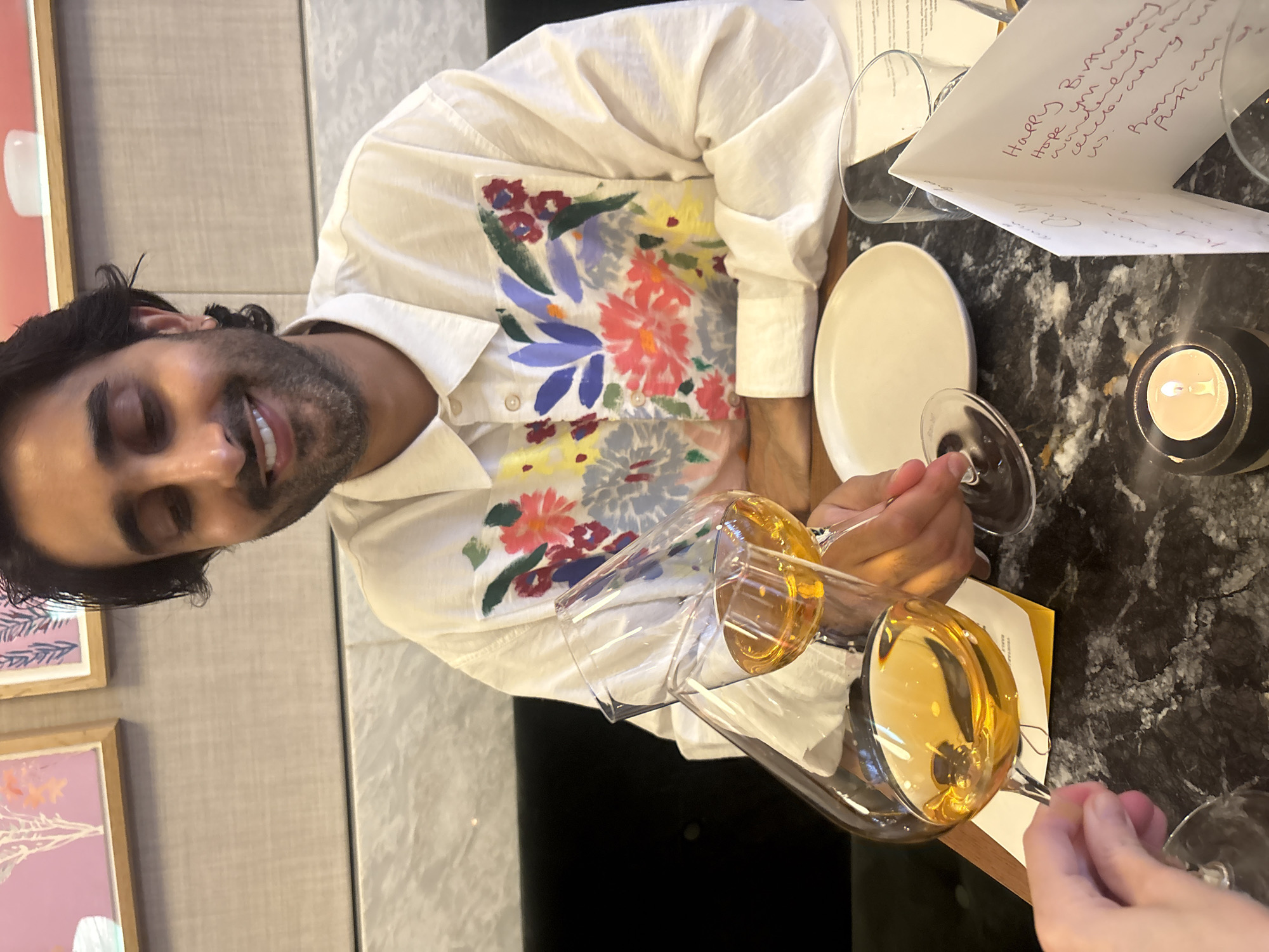 An overhead shot shows a person seated at a table in what appears to be a restaurant, raising a glass of amber liquid in a toast. The individual, a man with dark hair and a beard, is wearing a white button-down shirt with a colorful floral print. He is smiling and looking down, focused on the toast. Two wine glasses are visible, one in the man's hand and another being held by an unseen person whose hand is in the lower left corner of the frame. The table surface is black marble, and a white plate and a lit candle are also present. A folded card with handwritten text is visible in the foreground. The background shows a wall with framed artwork and a hint of cushioned seating, suggesting an indoor dining setting. The overall atmosphere suggests a celebratory occasion.