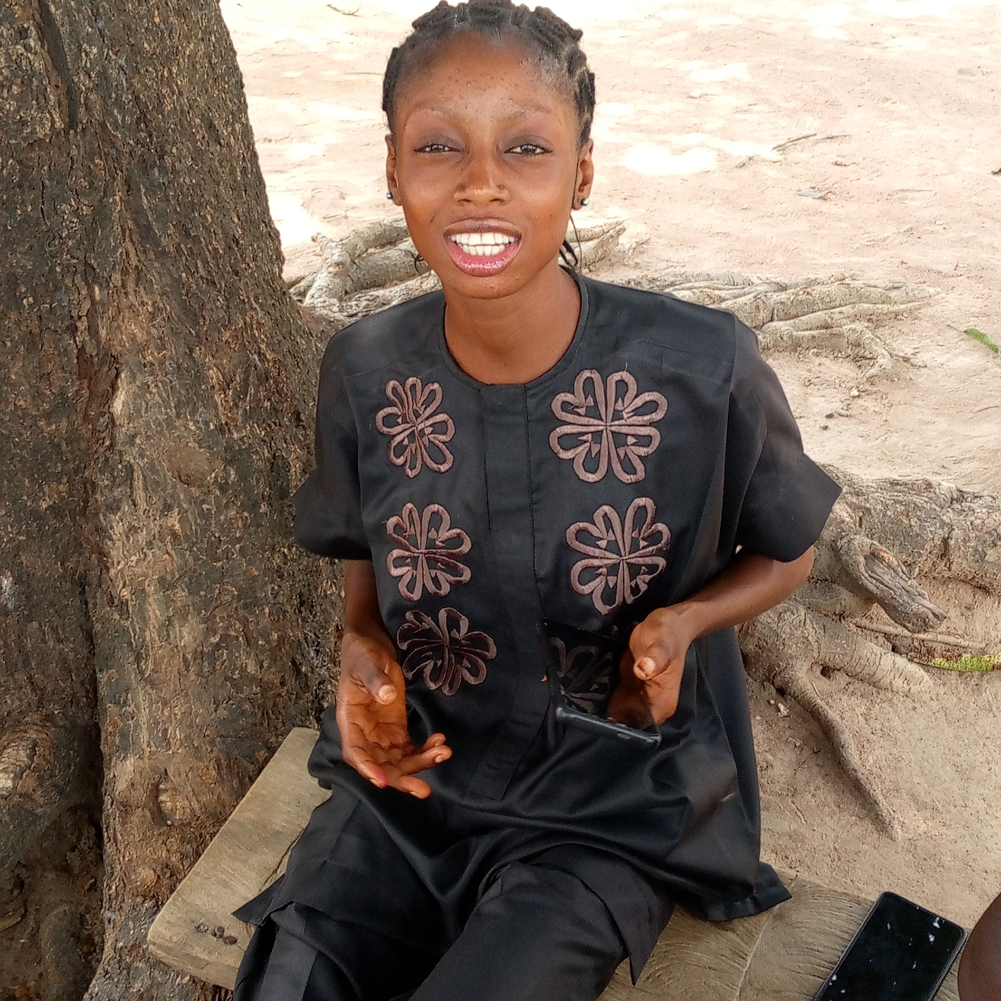 A young woman sits outside near a tree in Mararaba, Nigeria. She is wearing a black short-sleeved top with brown flower-like designs and black pants. She holds a black smartphone in her hands, while another lies on the wooden bench next to her. The ground is sandy and roots are visible.