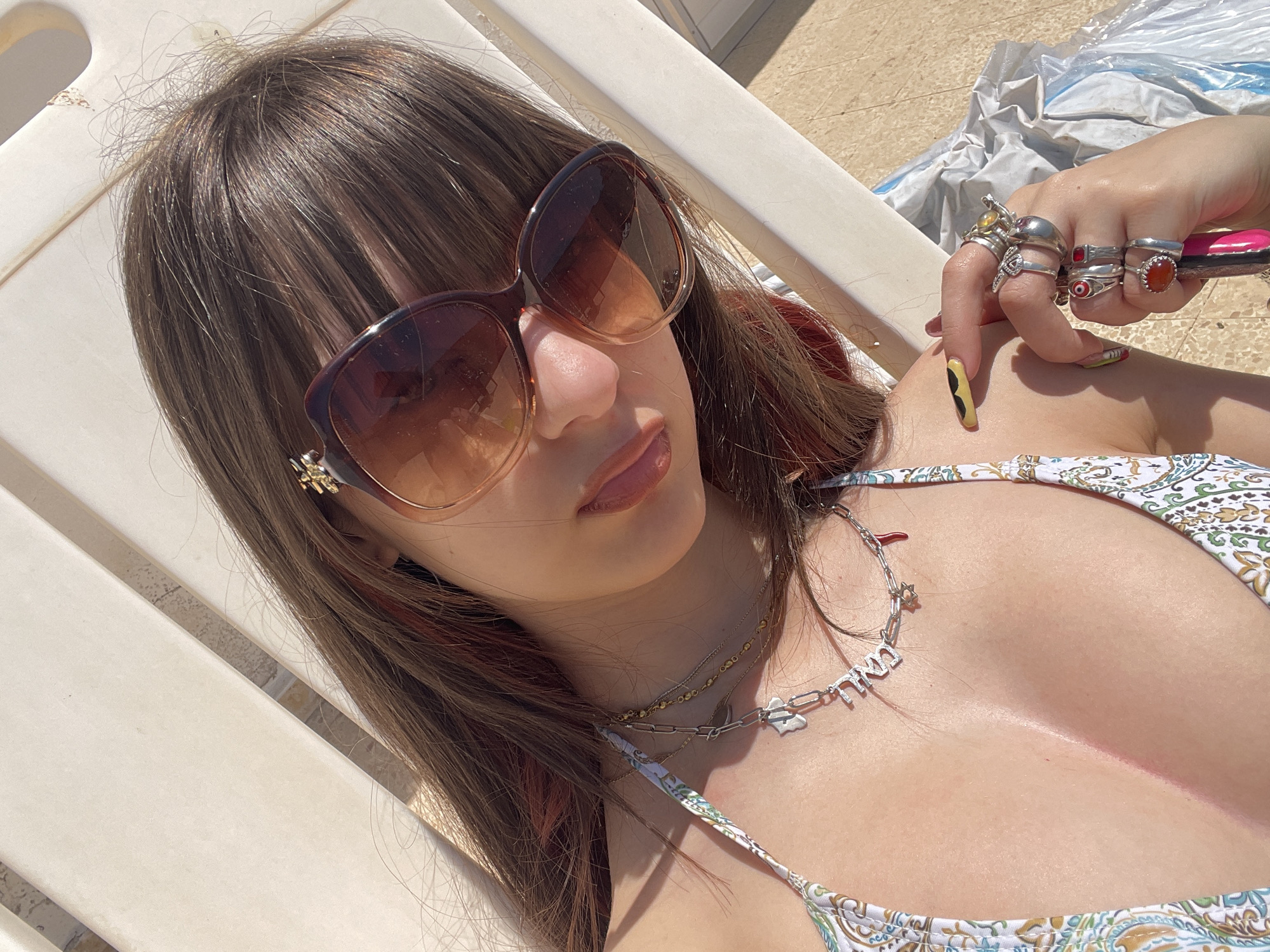 A young woman is reclining on a white chair outdoors in bright sunlight. She is wearing brown sunglasses and a patterned bikini. She has light brown hair with red highlights and is wearing multiple necklaces. Her hand is raised near her shoulder and is adorned with several rings. Her nails are painted with a unique yellow and black design. The location is likely in Petah Tikva, Israel, as indicated by the prompt's request, although no specific landmarks are visible in the image to confirm this location.
